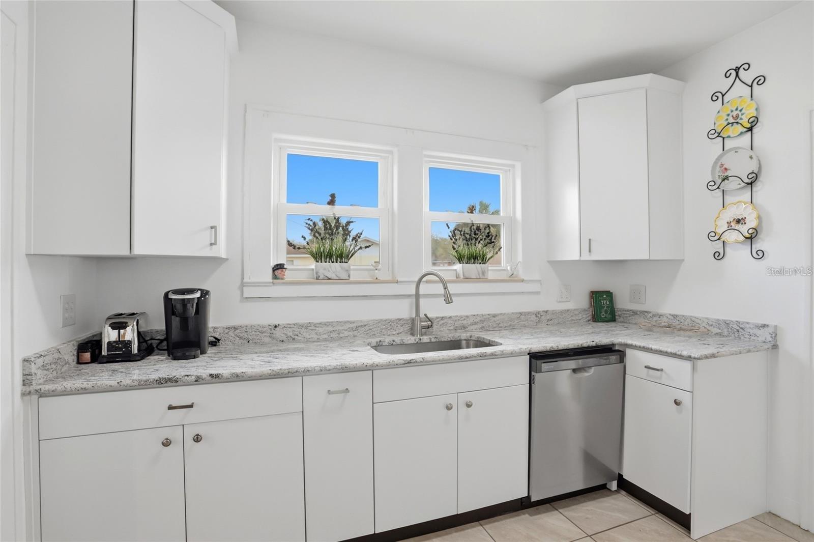 Bright white kitchen with granite counters, a stainless-steel dishwasher, and a sink beneath two windows.