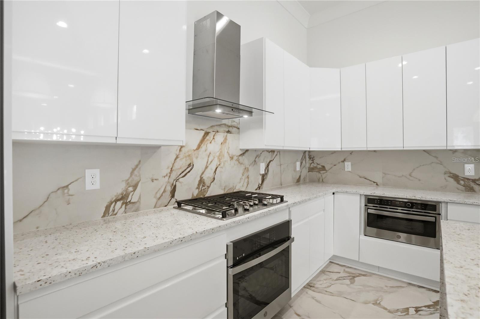 Modern white kitchen with a gas cooktop, veined stone backsplash, and built-in stainless ovens.