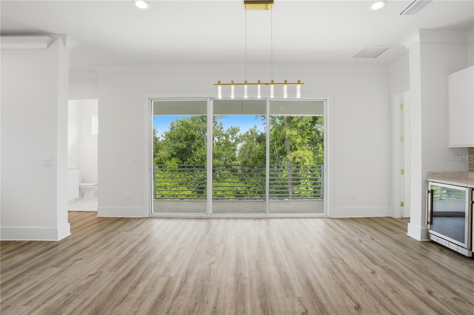 Vacant interior with wood-look floors, sliding doors to a tree-lined balcony, and a modern pendant light.