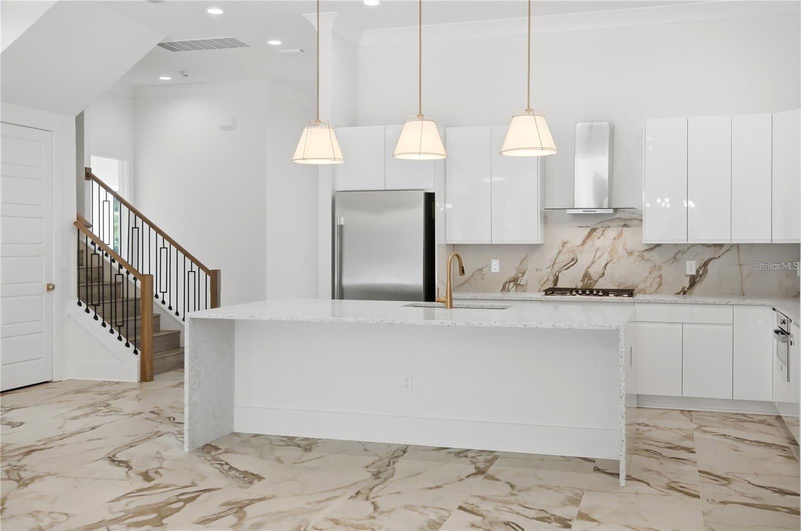 Modern white kitchen with a large island, marble-patterned surfaces, gold fixtures, and an adjoining staircase.