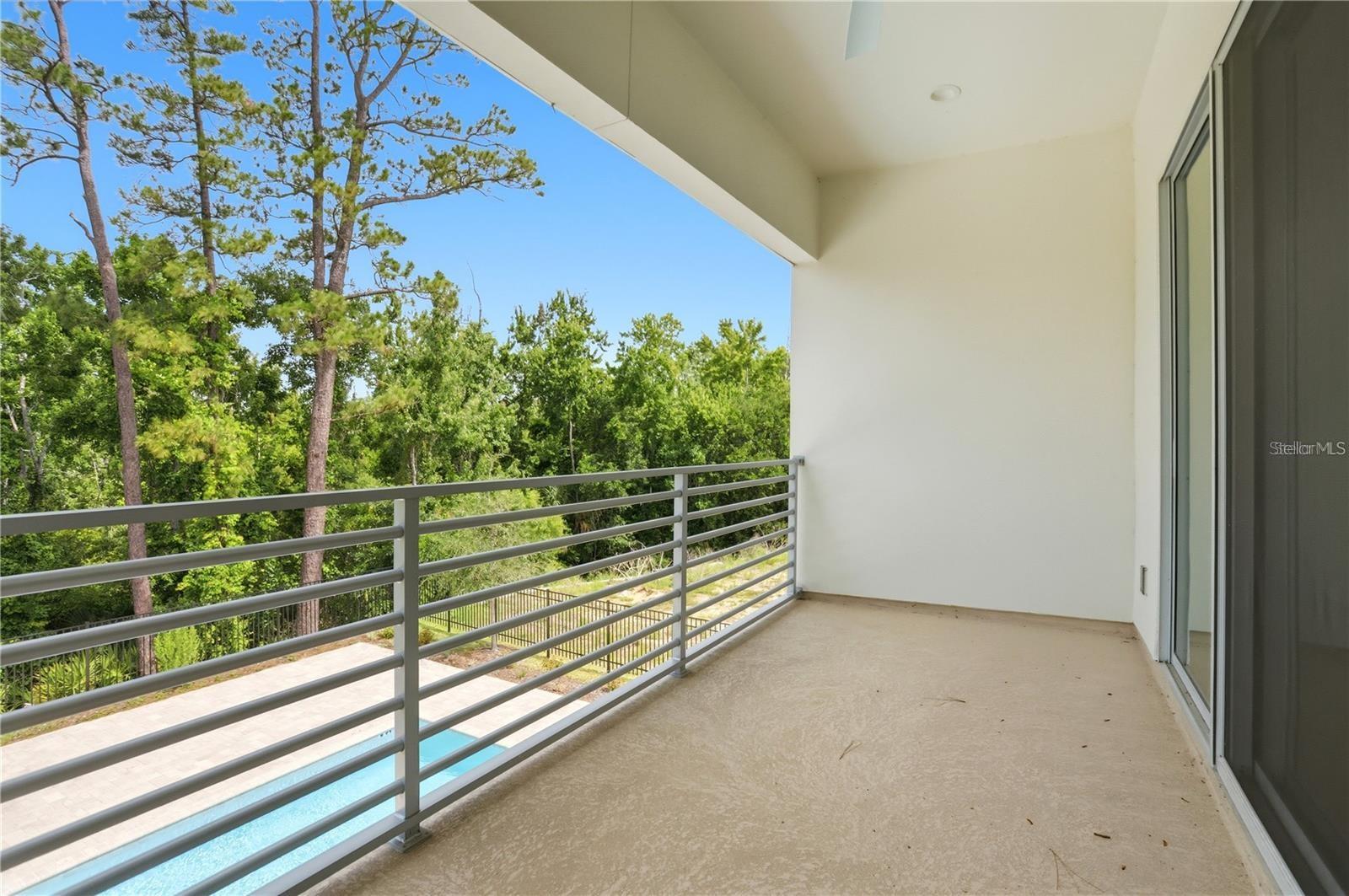 Covered balcony with gray railing overlooking trees and a glimpse of a swimming pool.