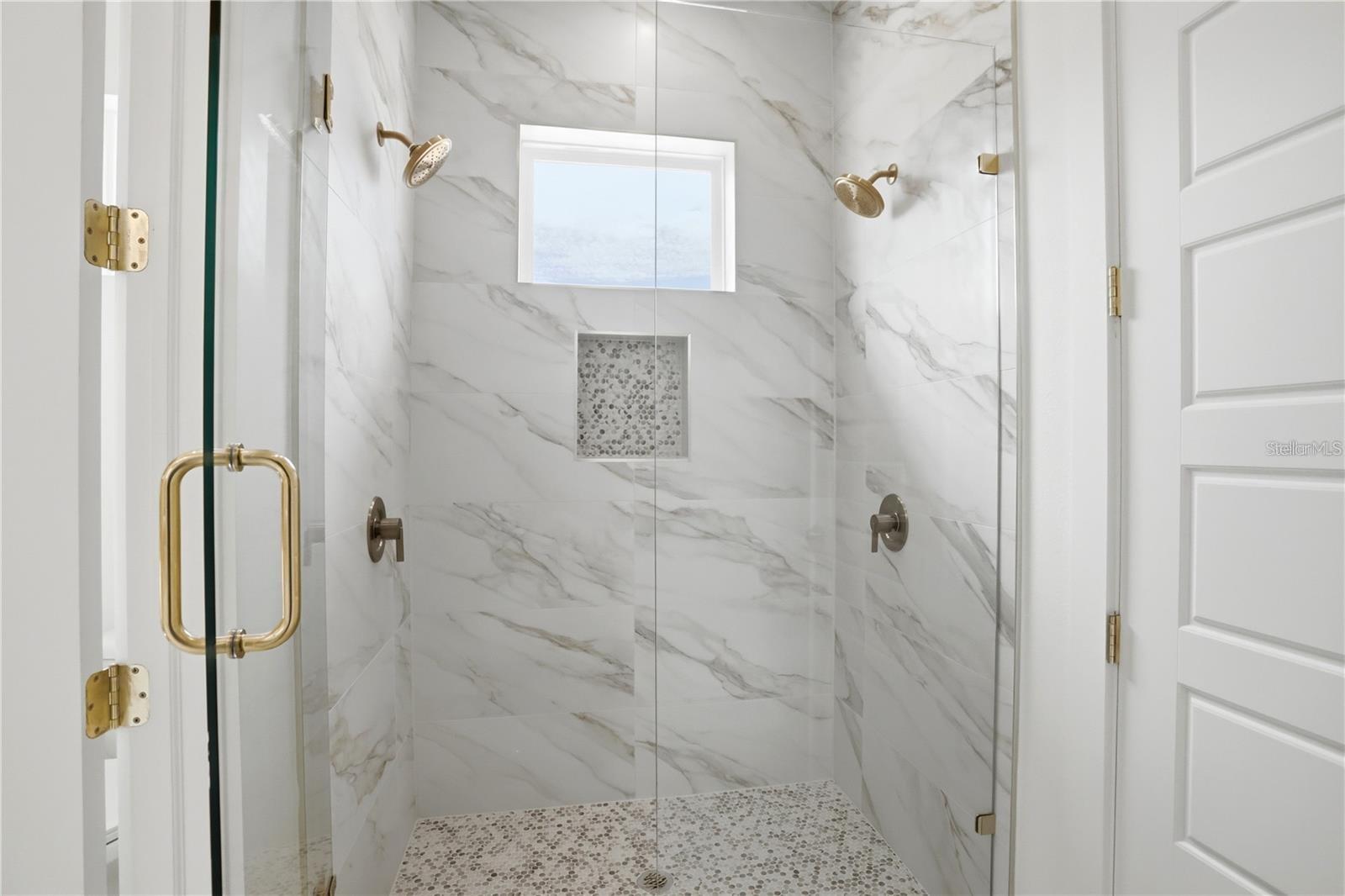 Walk-in shower with marble-look tile, brass showerheads, a window, and pebble flooring.