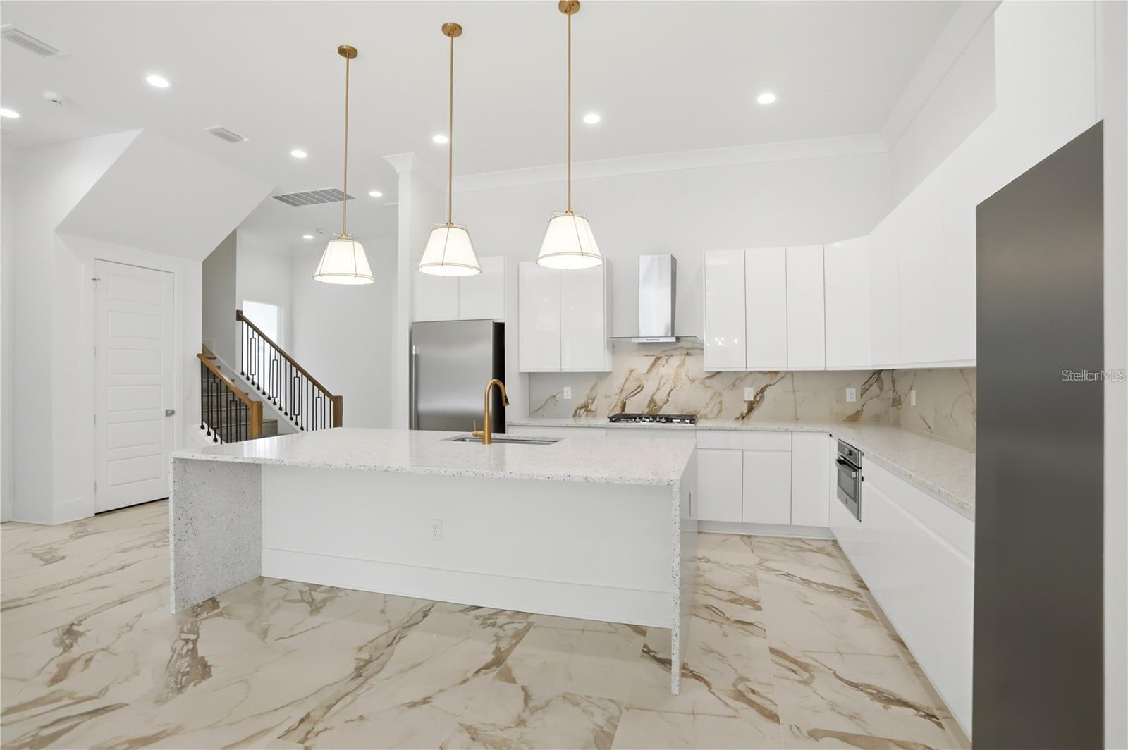 Modern white kitchen with a large island, gold faucet, marble-look surfaces, and three pendant lights.
