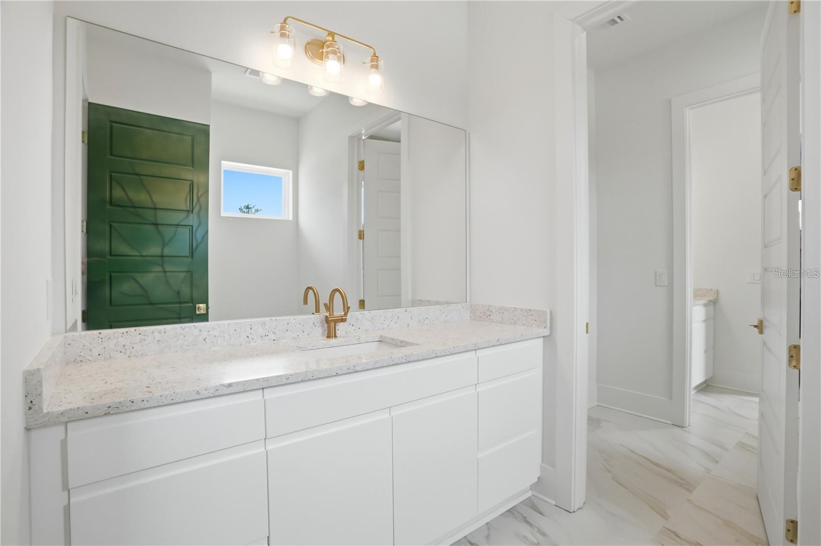 Bright bathroom vanity with a speckled stone counter, gold faucet, and mirror reflecting a green door.