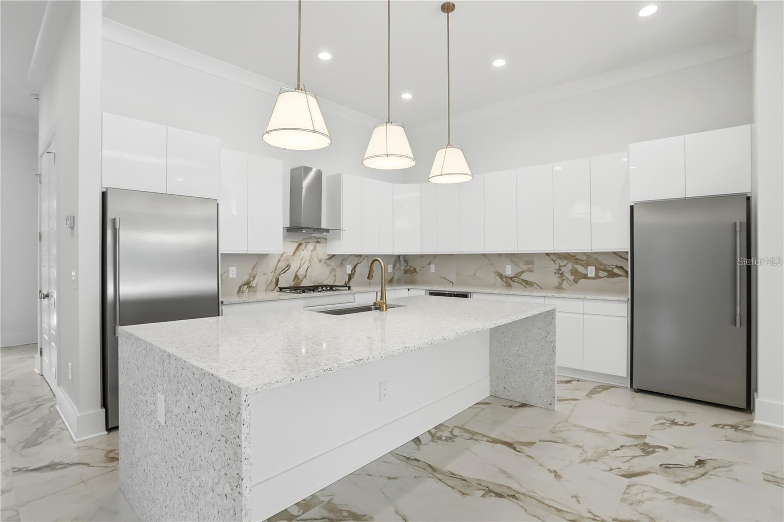 Modern white kitchen with a large stone island, stainless steel refrigerators, and veined marble-look surfaces.
