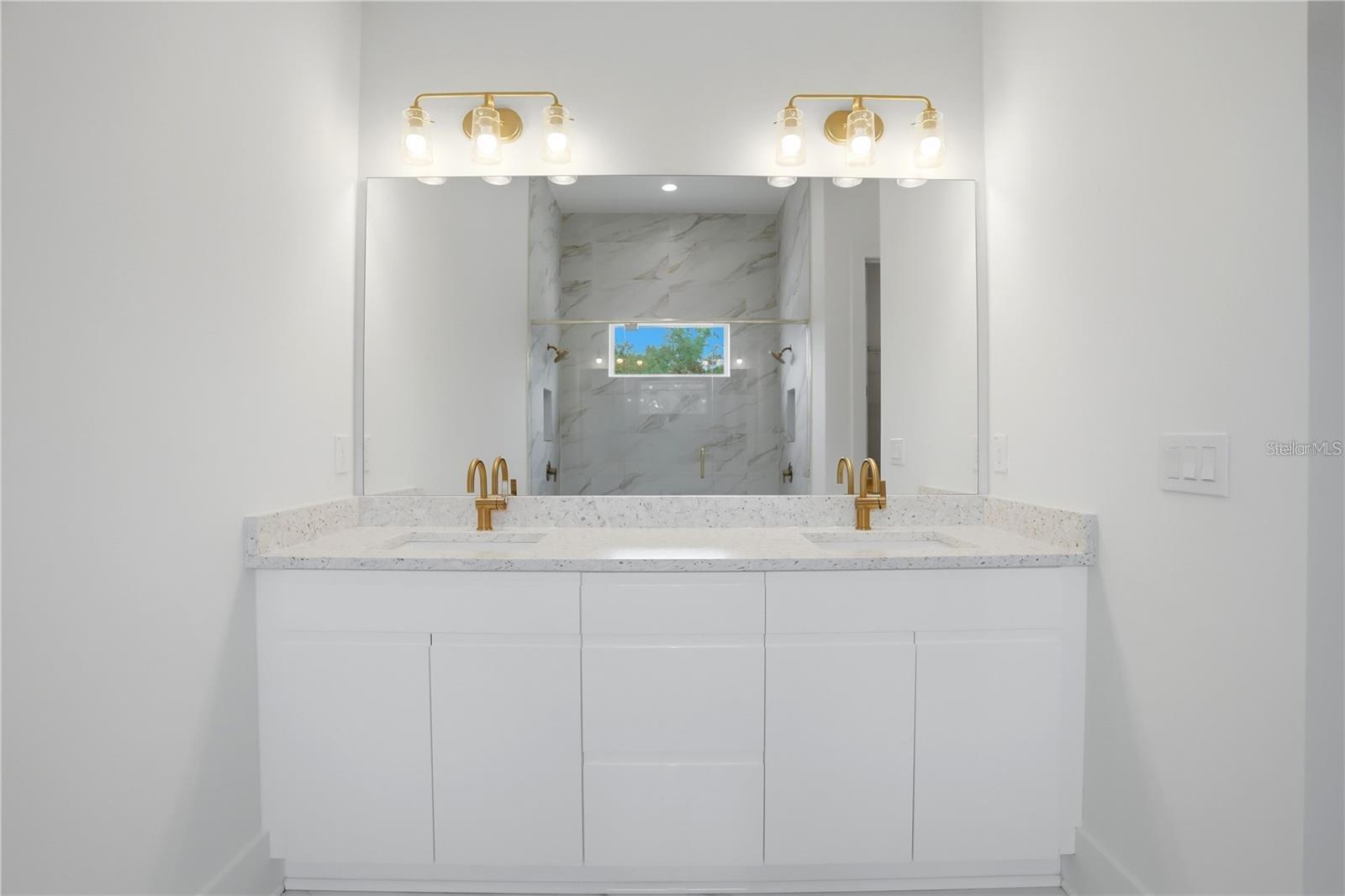 Bathroom vanity with gold faucets and a large mirror reflecting a marble-look shower.