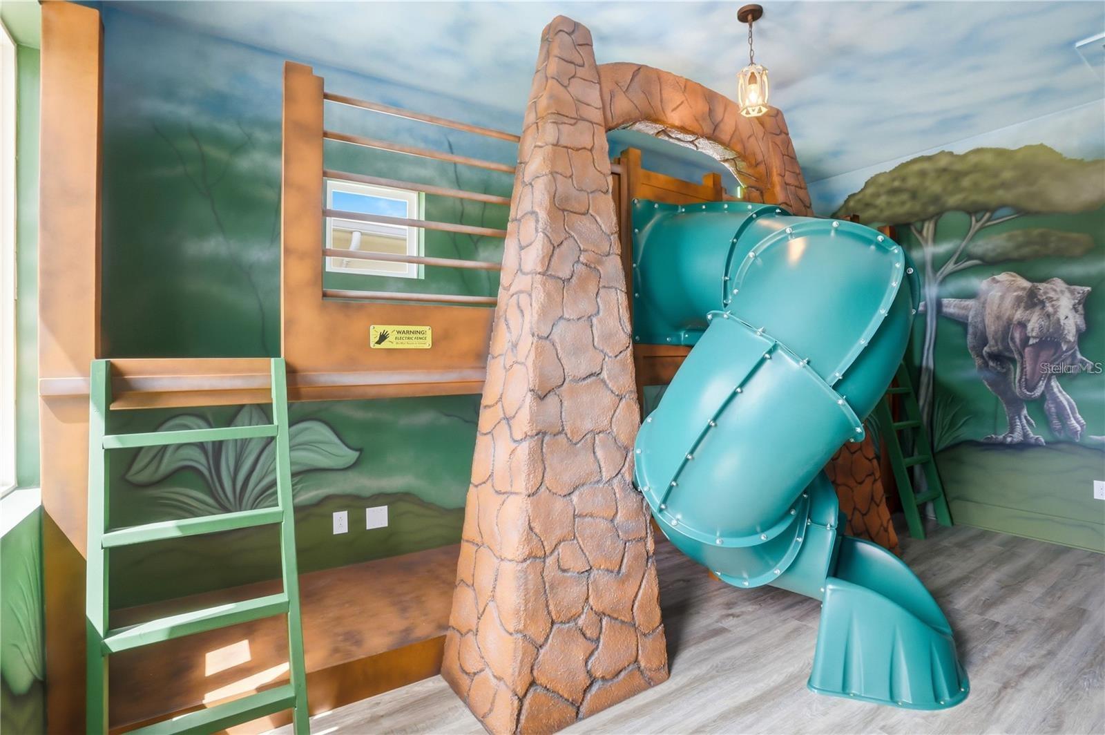 Children’s playroom with a teal tube slide, faux-stone tower, green ladder, and dinosaur mural.