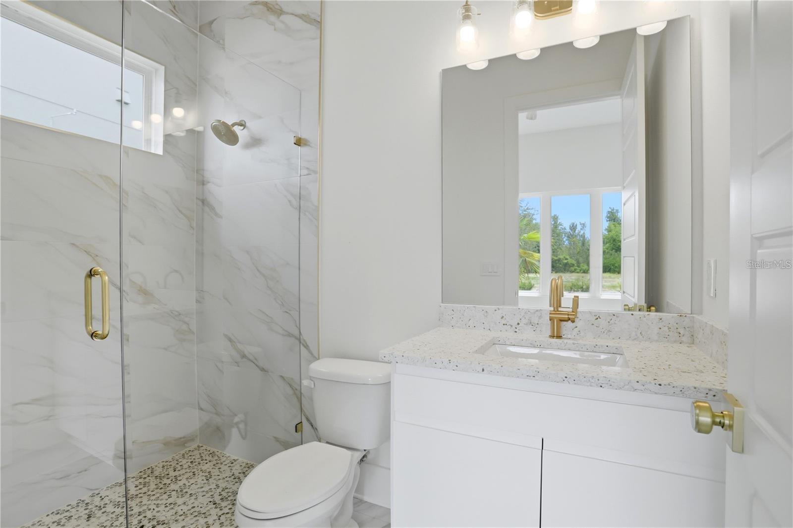 Modern bathroom with marble-look shower tile, pebble flooring, and a white vanity with gold fixtures.
