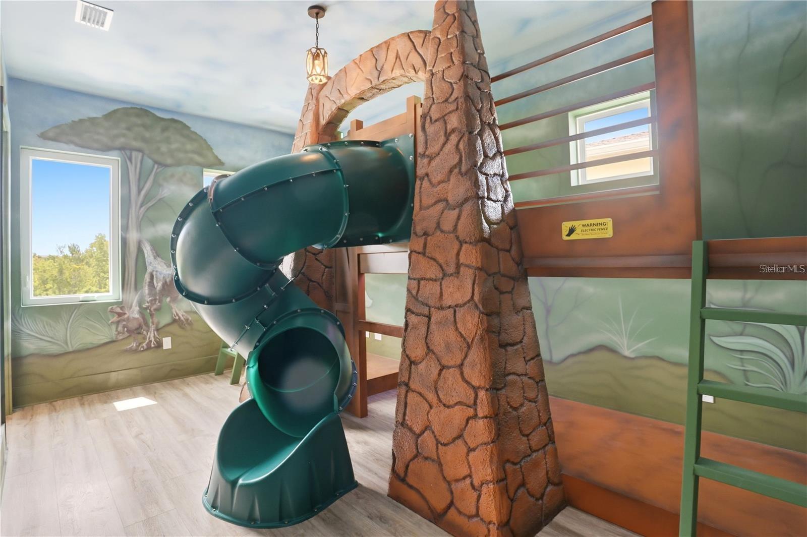 Indoor playroom with a green spiral slide, faux stone tower, wooden climbing platforms, and woodland murals.