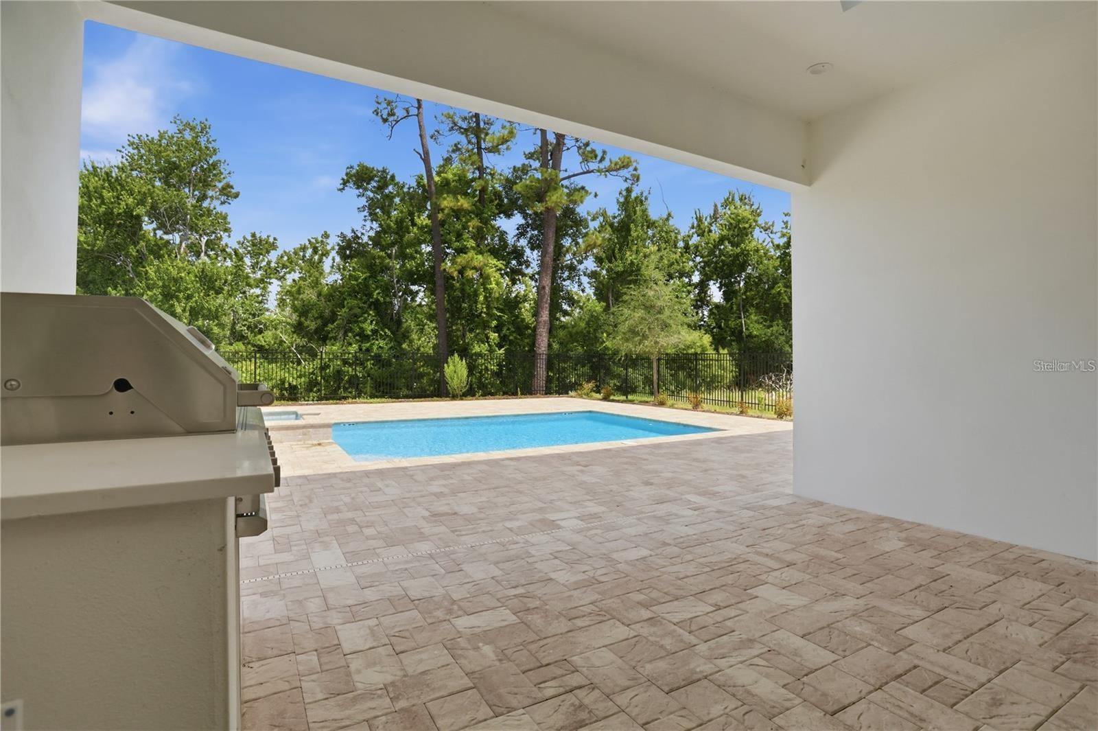 Covered patio with built-in grill and paver deck overlooking a fenced pool and mature trees.