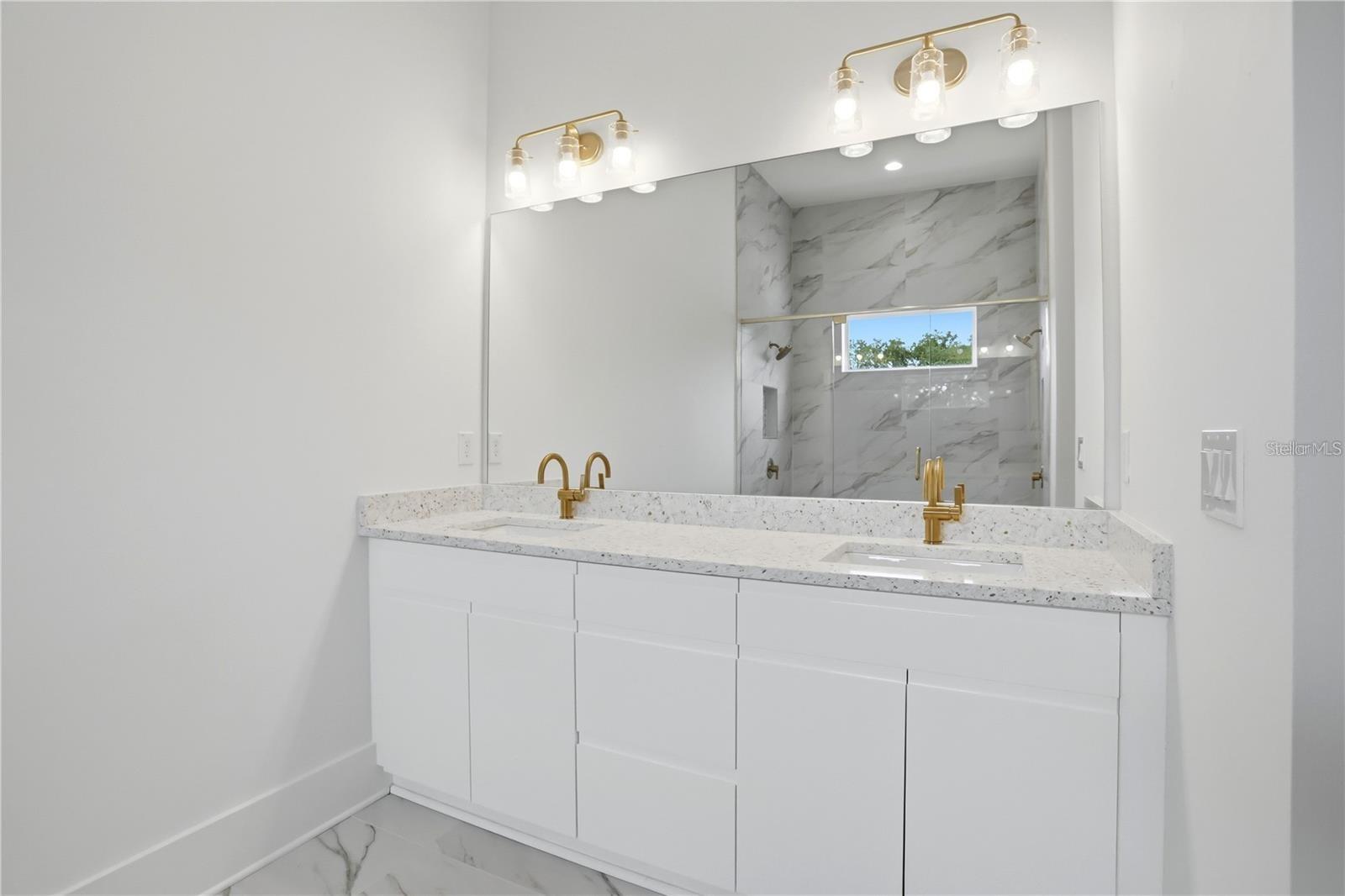 Bathroom with a double-sink white vanity and gold fixtures, facing a marble-look tiled shower.