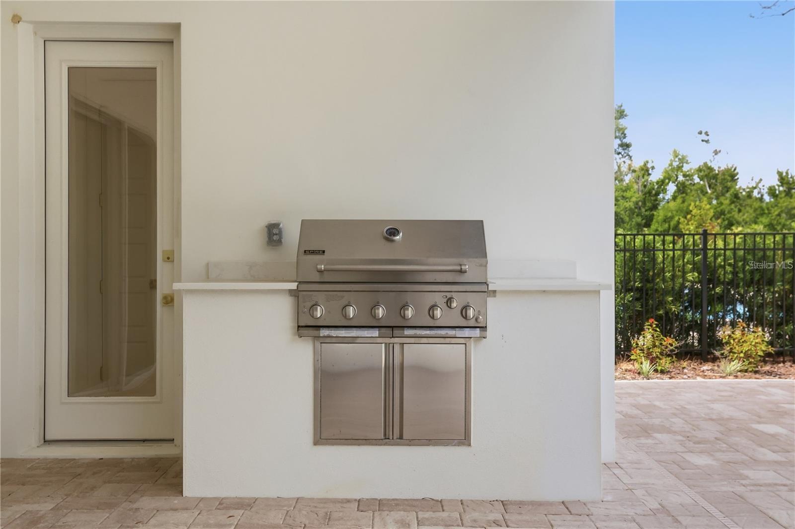 Built-in stainless steel grill at a covered outdoor kitchen beside a paved patio.