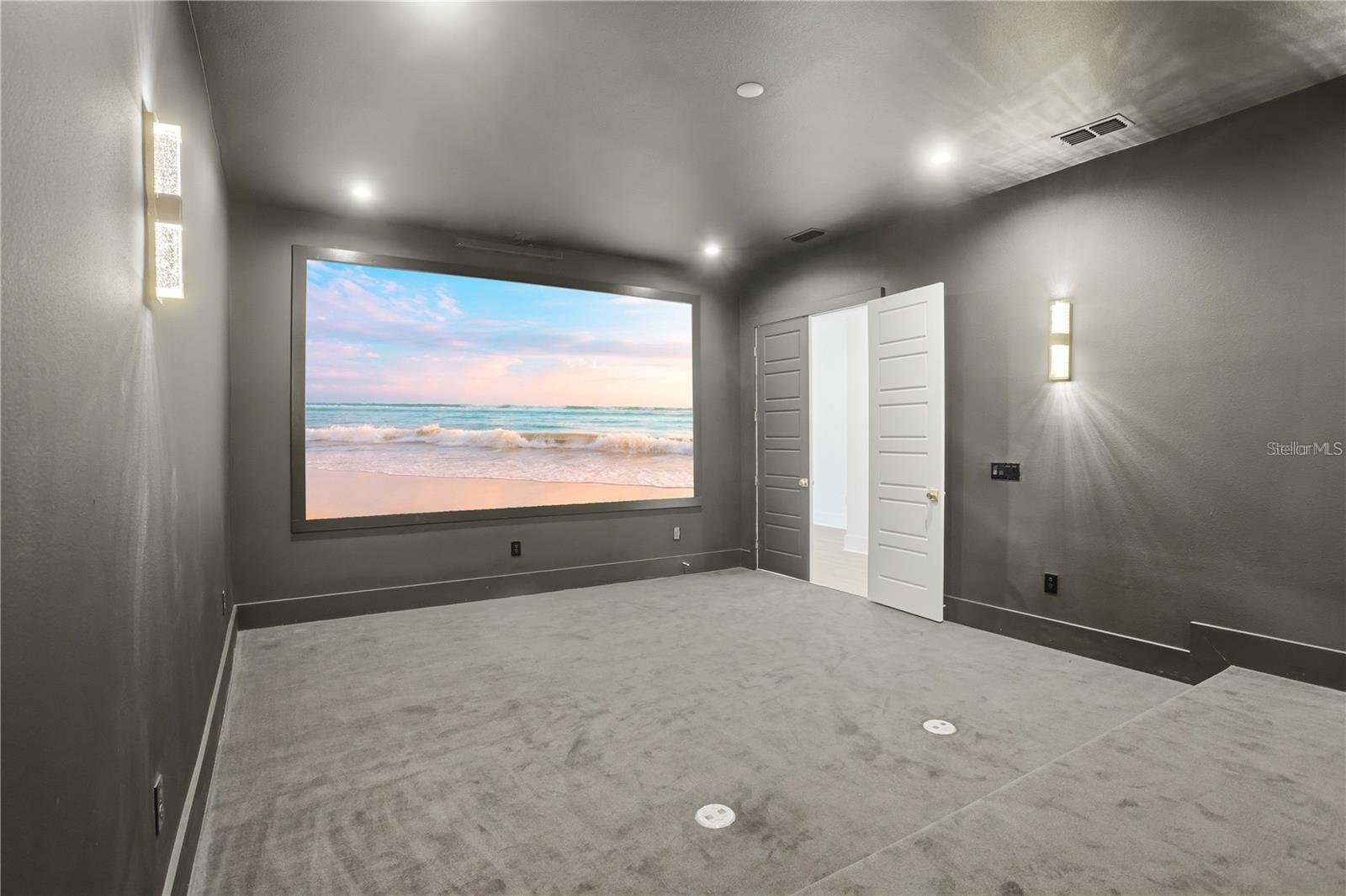 Empty home theater with gray carpeting and a large screen displaying a beach scene.