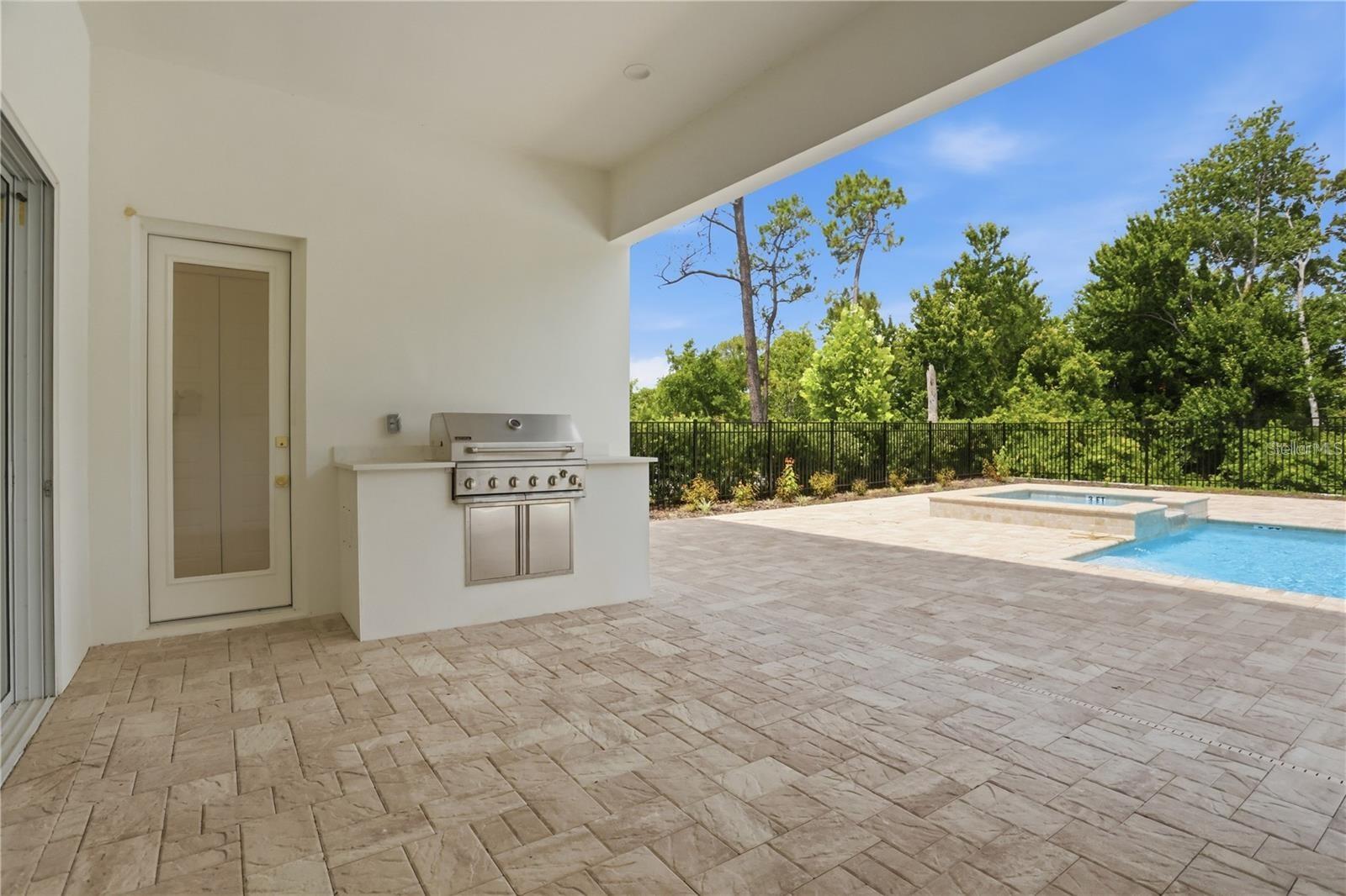 Covered patio with built-in grill overlooking a fenced pool and raised spa.