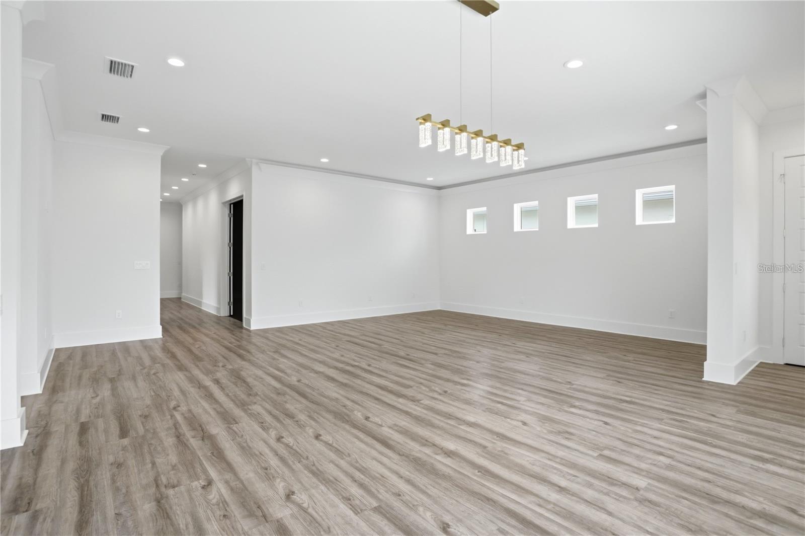 Empty open room with gray plank flooring, four high windows, and a modern gold chandelier.