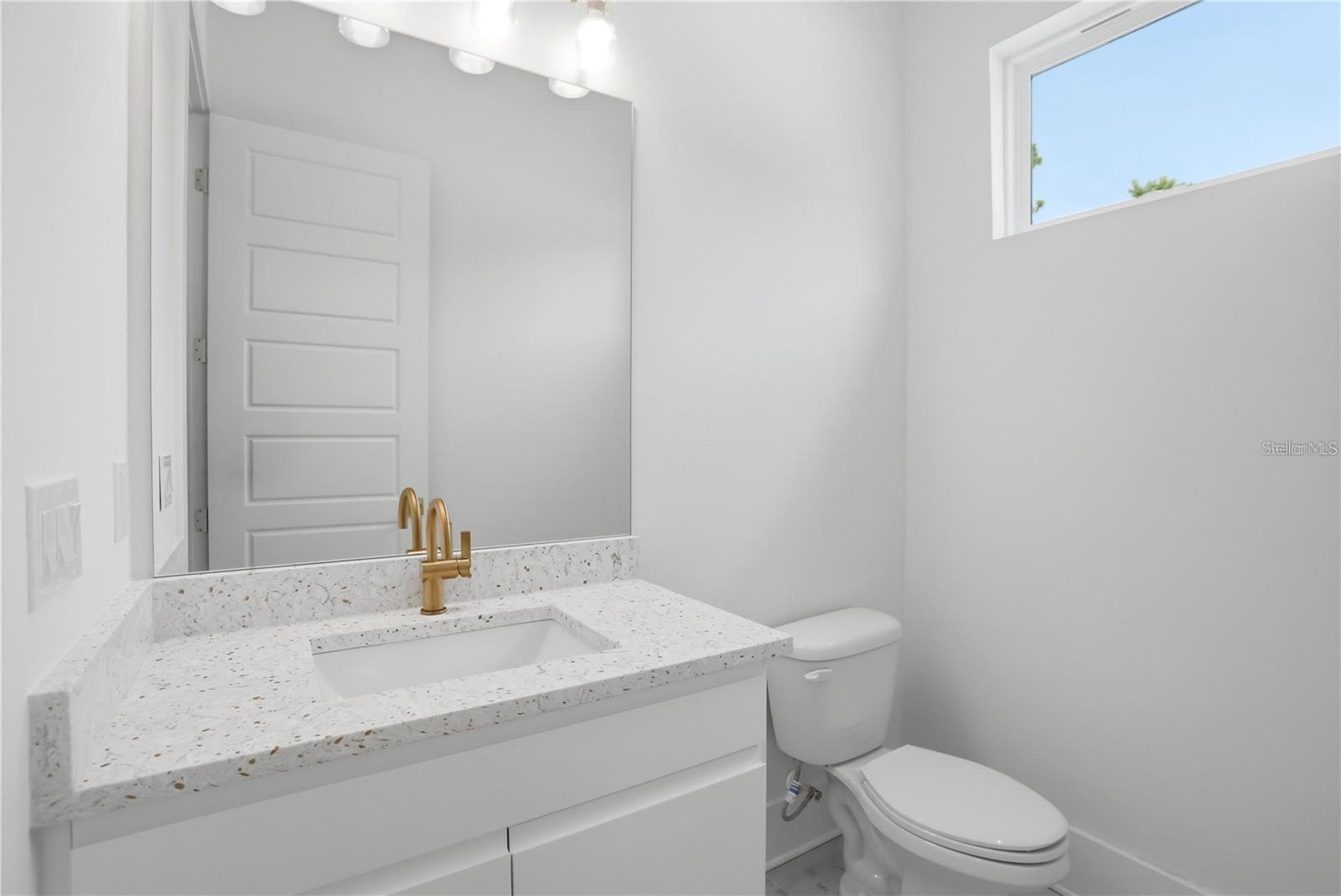 Bathroom with a speckled stone vanity, gold faucet, large mirror, and high window.