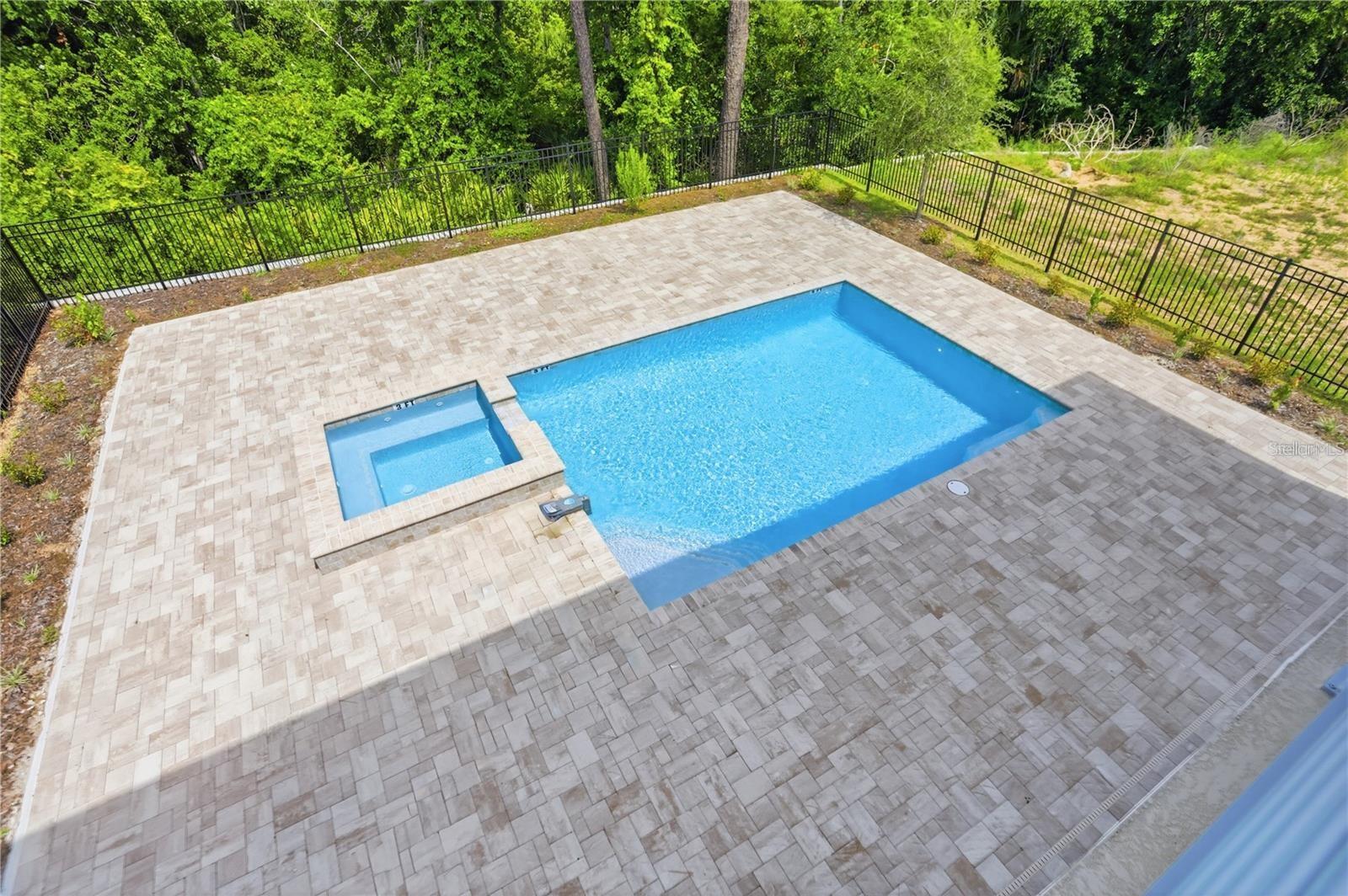 Aerial view of a blue swimming pool with an attached spa, broad paver patio, and wooded fence line.