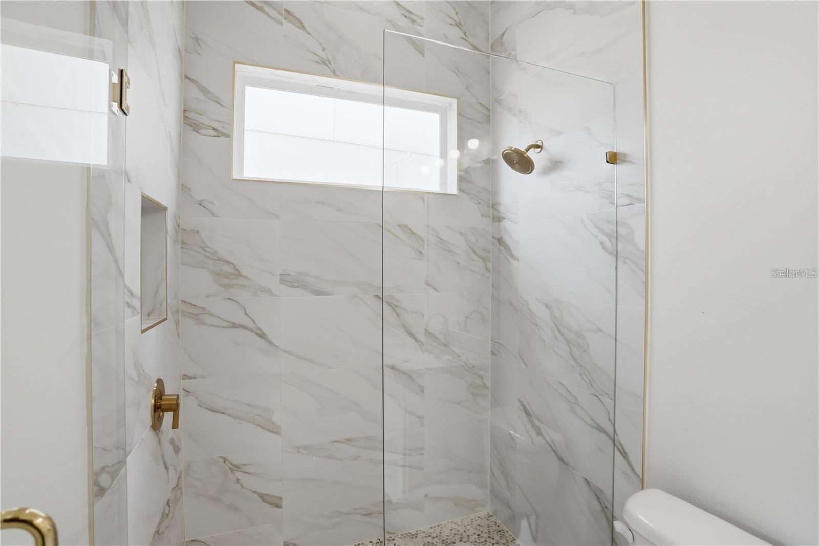 Marble-tiled bathroom shower with a glass enclosure, brass fixtures, and a high window.
