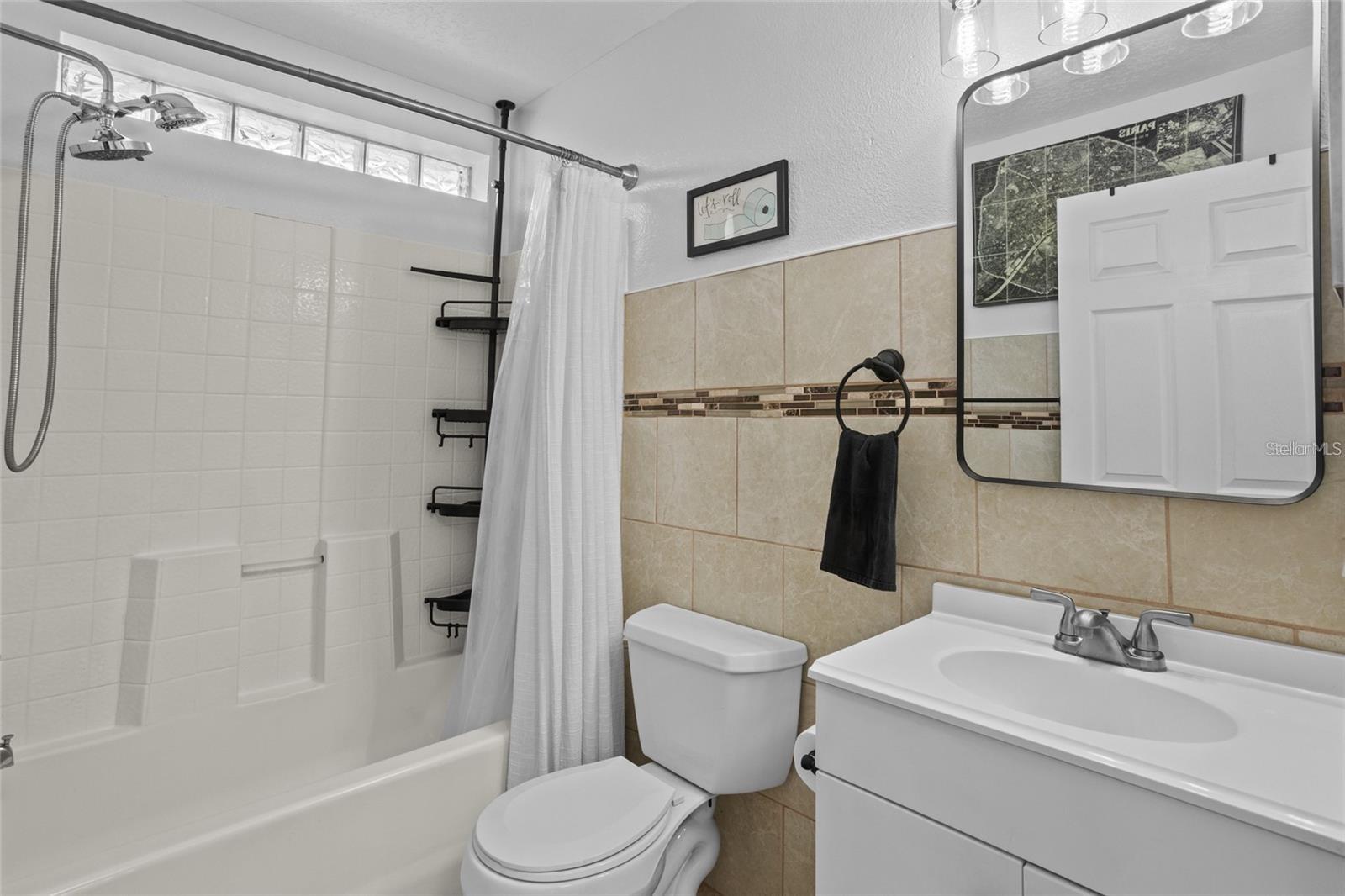 Bathroom with a white tub, shower curtain, toilet, and vanity against beige tiled walls.
