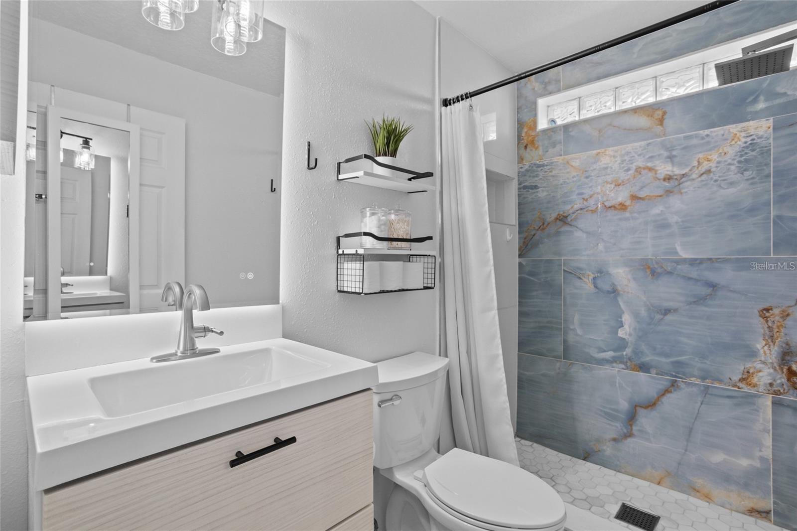 Updated bathroom with a white vanity and toilet beside a blue-gray marble-look tiled shower.