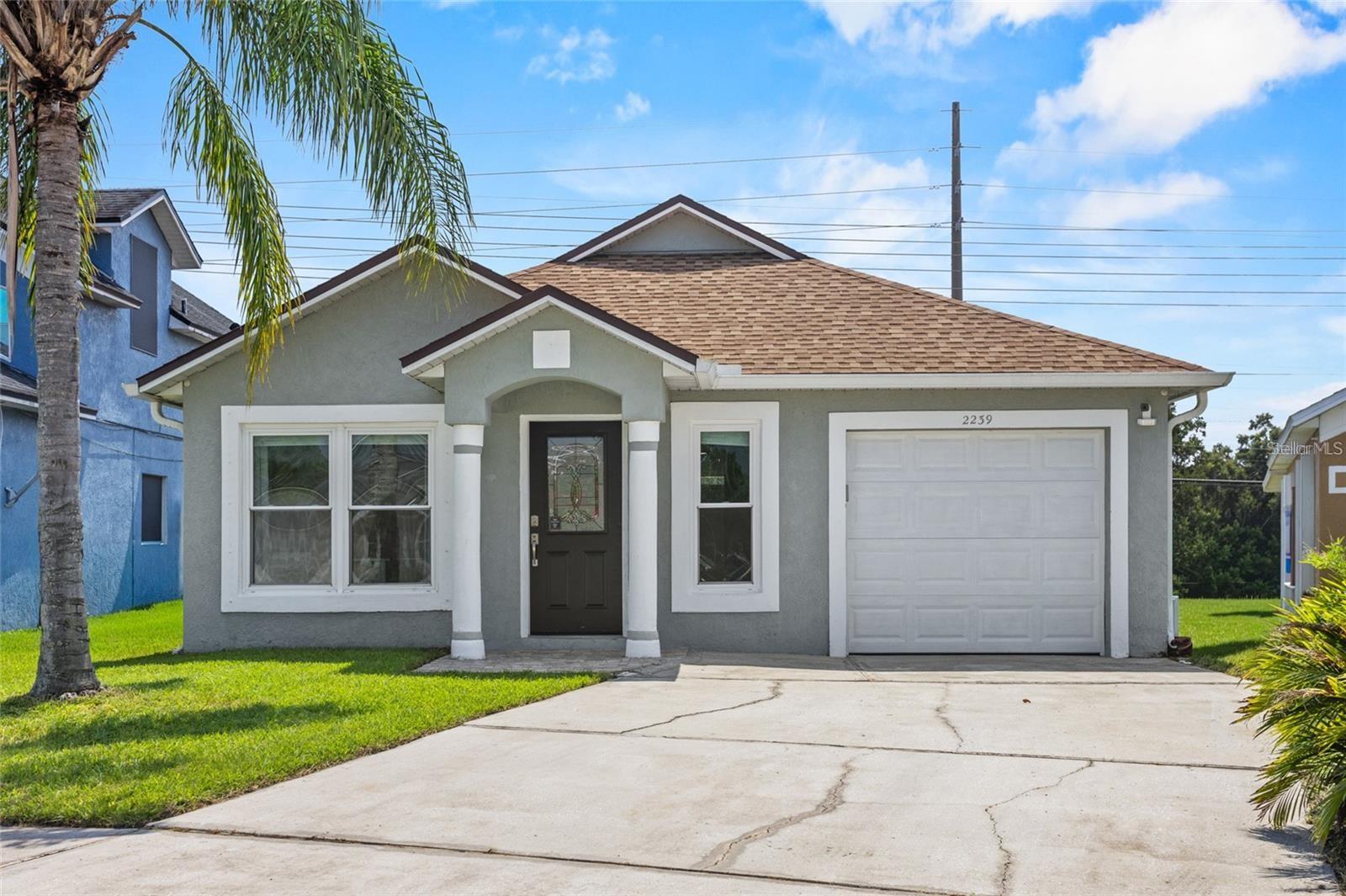 Gray single-story home with an attached garage, palm tree, and wide concrete driveway.