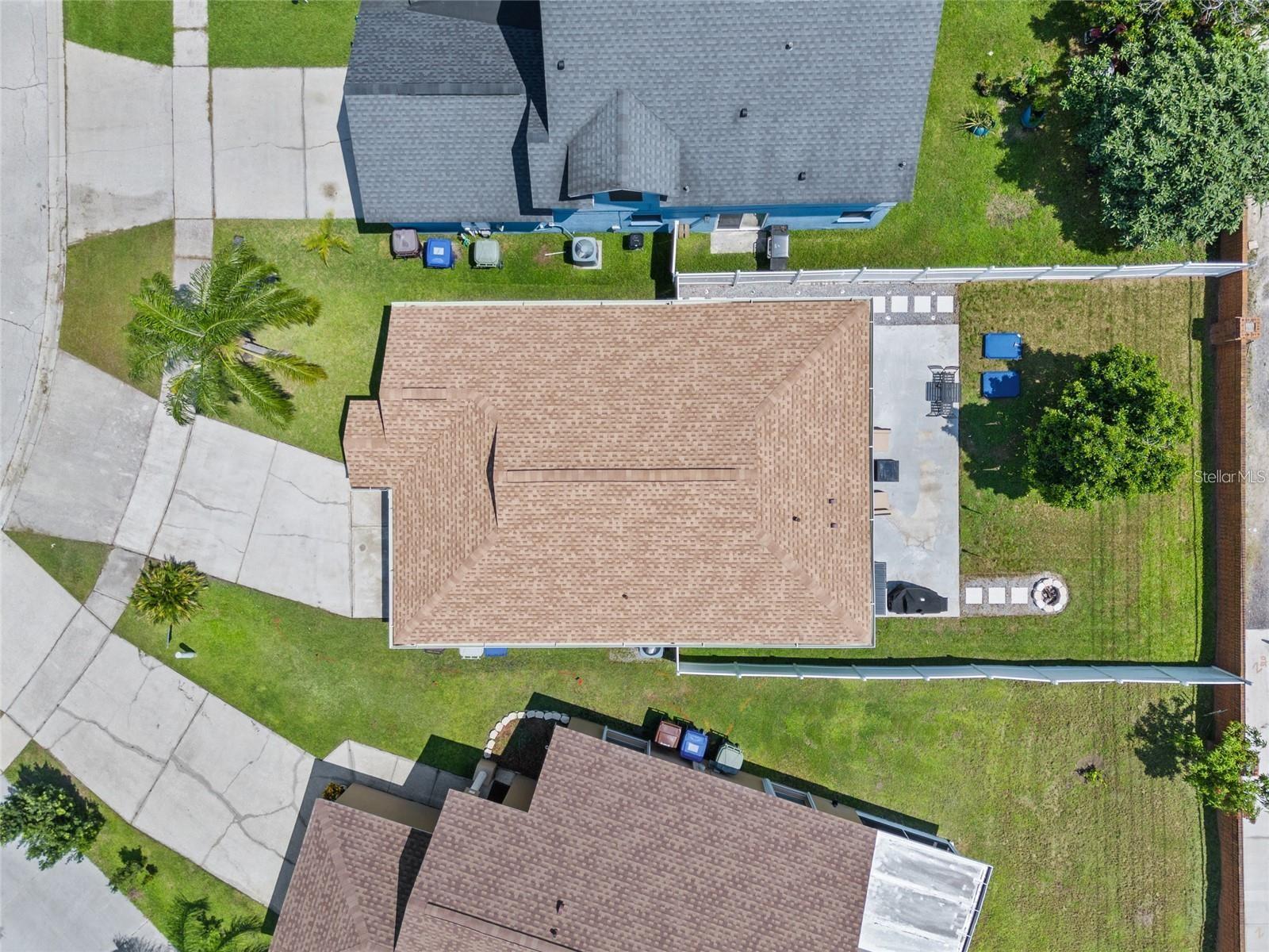 Aerial view of a tan-roofed home, fenced yard, driveway, and concrete patio.