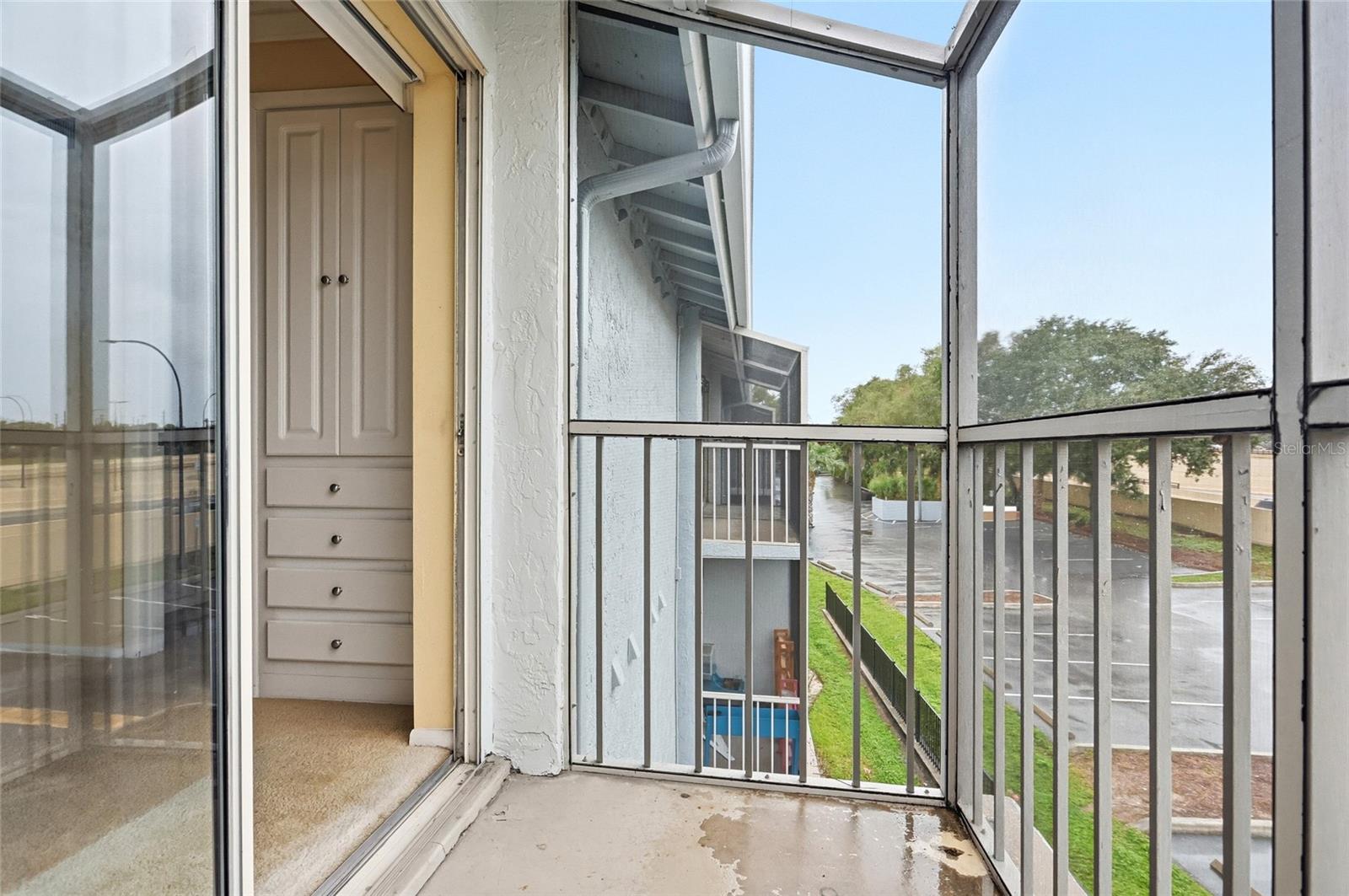 Screened balcony with white railing and concrete floor overlooking a parking area and neighboring buildings.