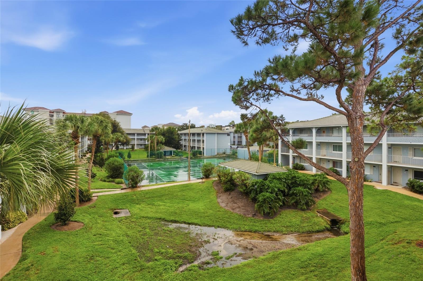 Condominium courtyard with a fenced tennis court, landscaped lawns, and multi-story buildings.