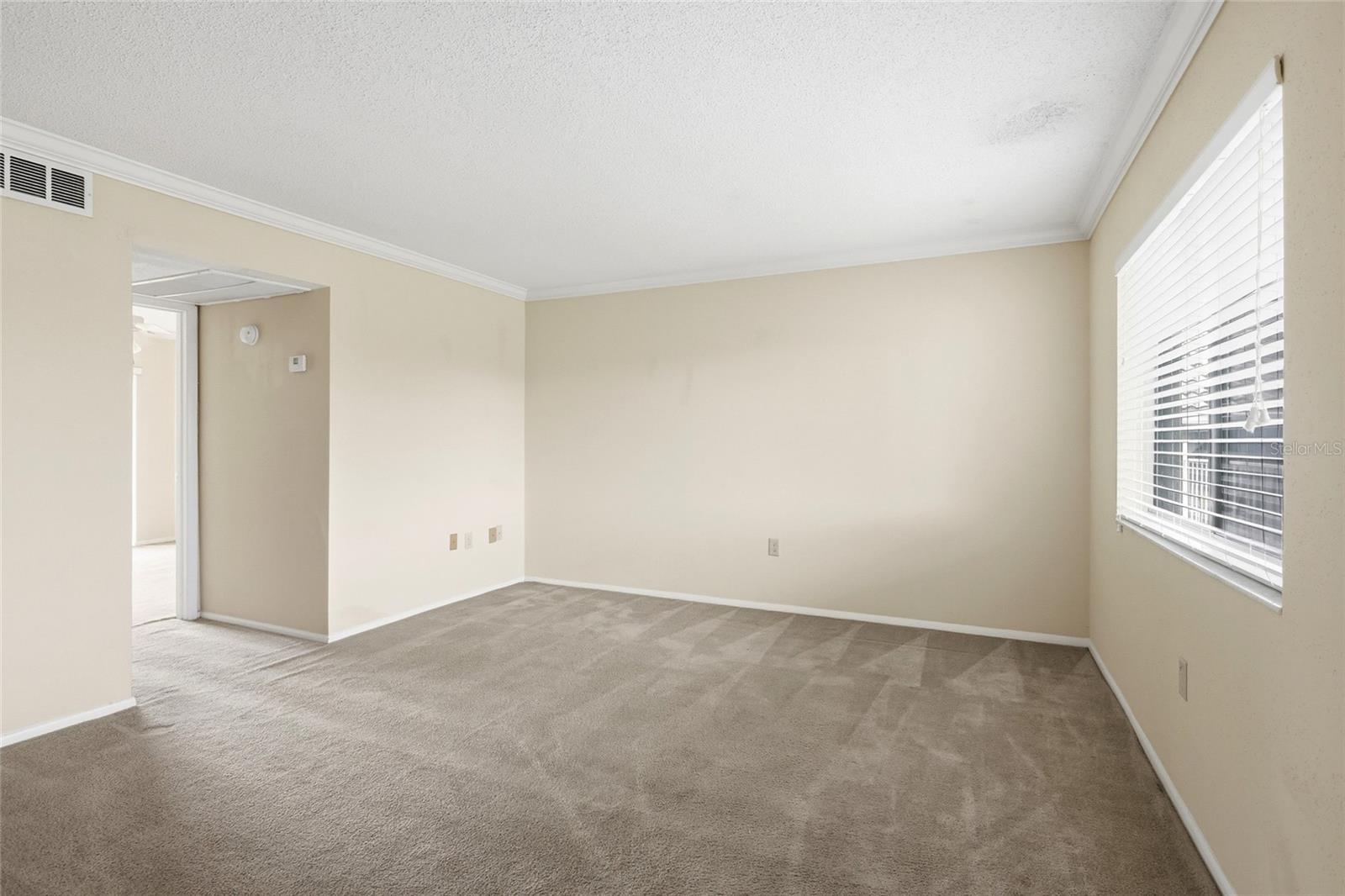 Empty carpeted room with beige walls, a wide window with white blinds, and an open doorway.