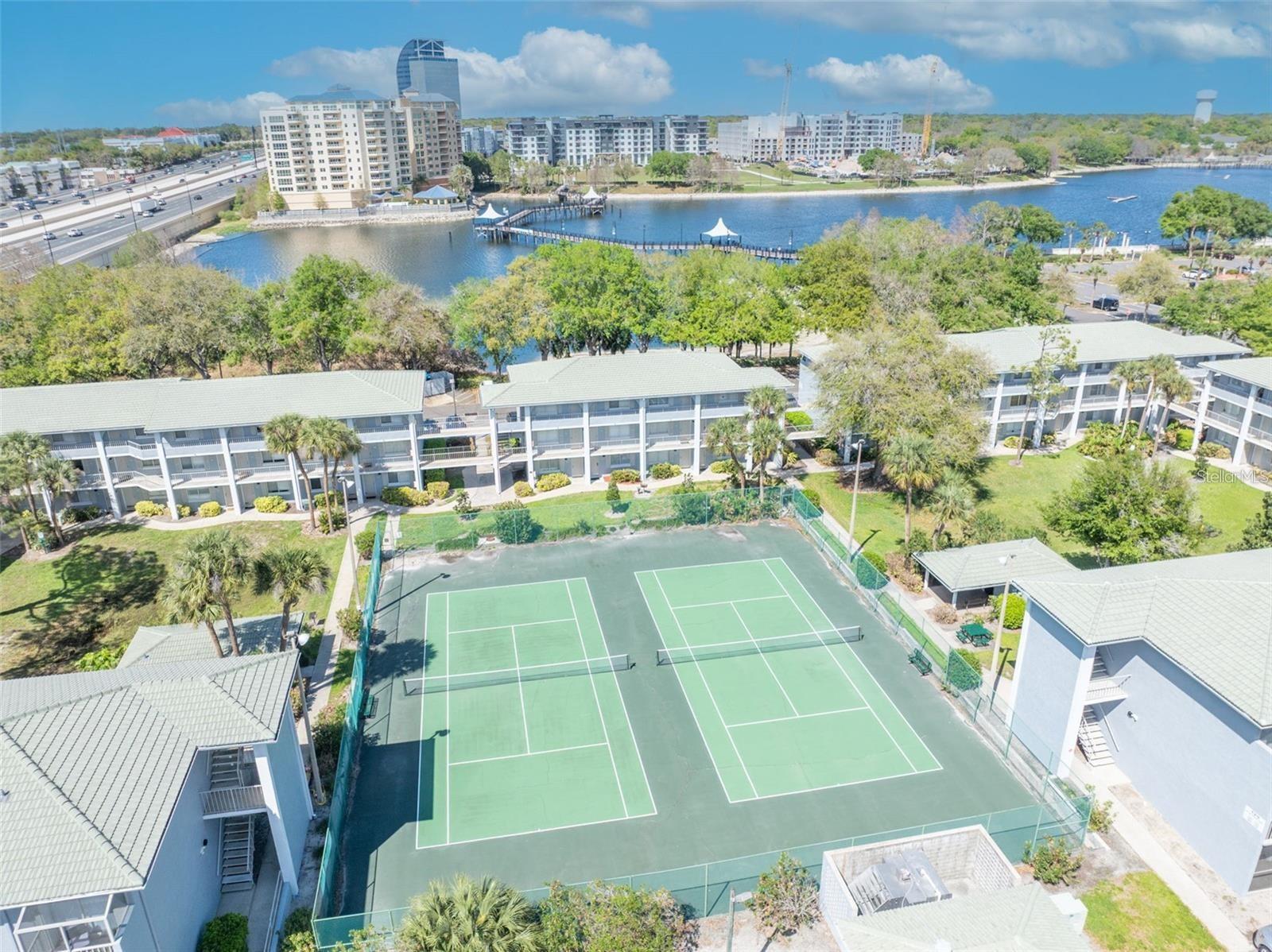 Aerial view of condominium tennis courts beside a lake, with a waterfront promenade and apartment buildings beyond.