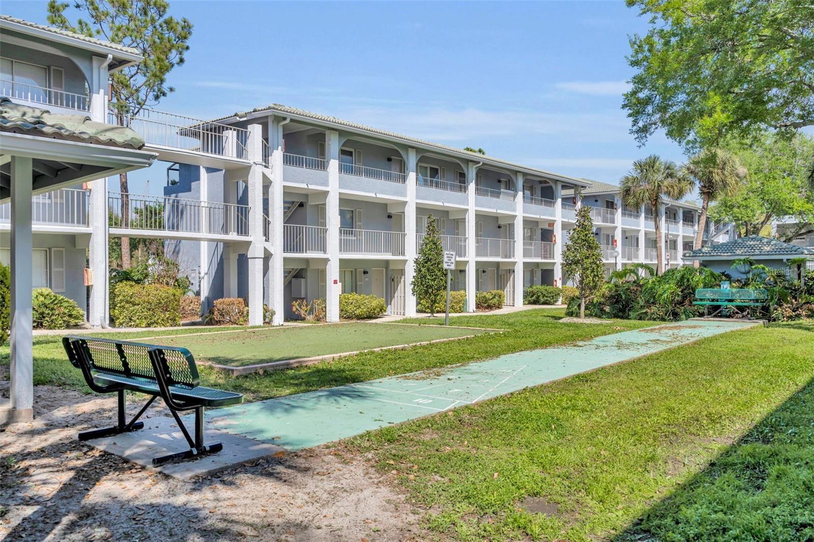 Condominium building with covered balconies beside a shuffleboard court and benches.