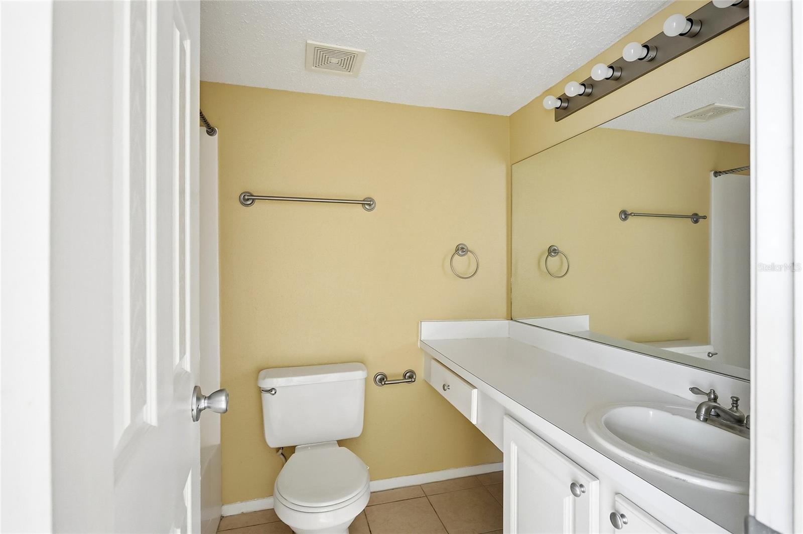 Bathroom with pale yellow walls, white vanity and toilet, large mirror, and tan tile floor.