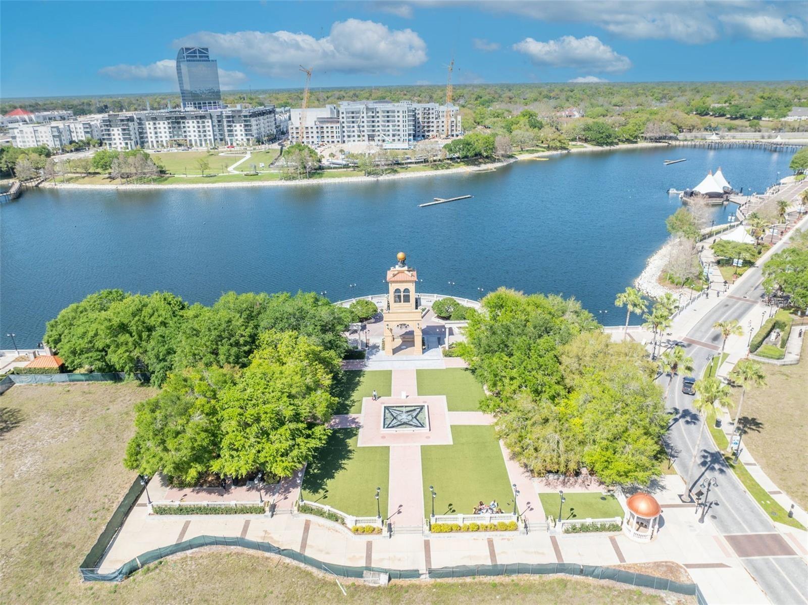 Aerial view of a lakefront park with a central monument and residential buildings across the water.