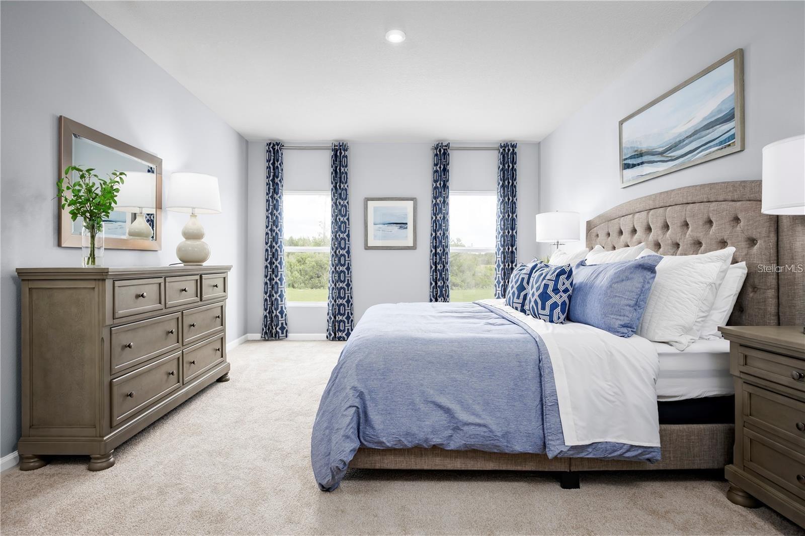 Staged bedroom with a tufted bed, blue-and-white bedding, gray dresser, and two curtained windows.