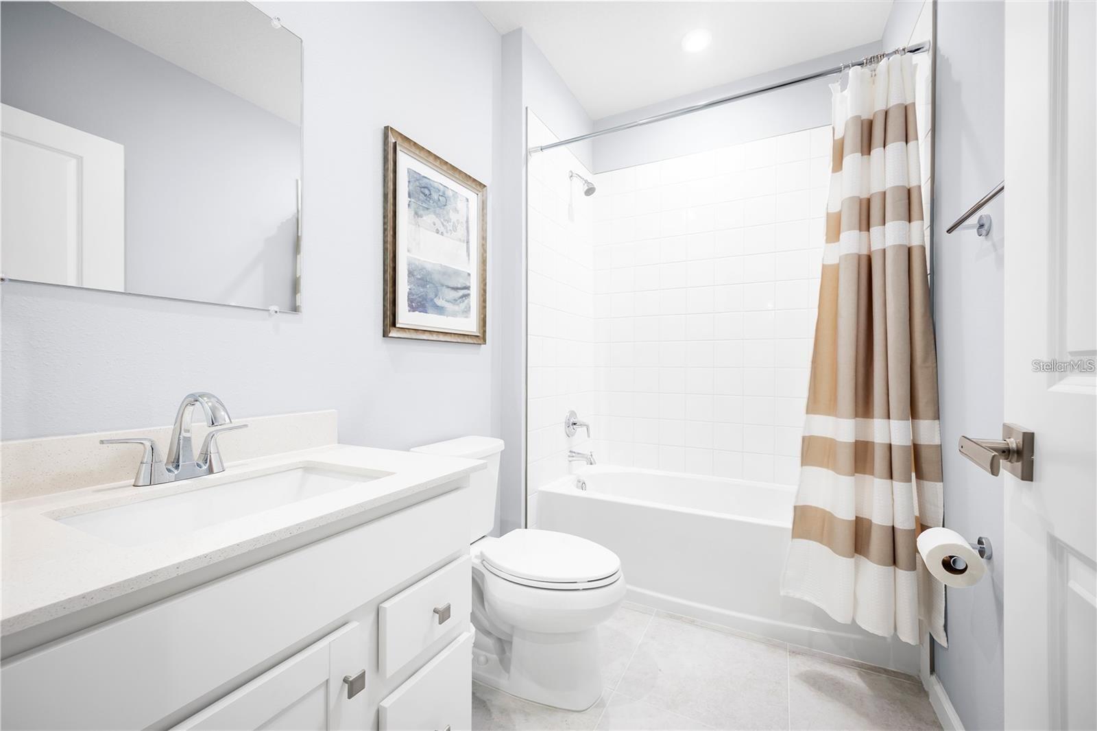 Bright bathroom with white vanity, toilet, and tub behind a tan-and-white striped curtain.