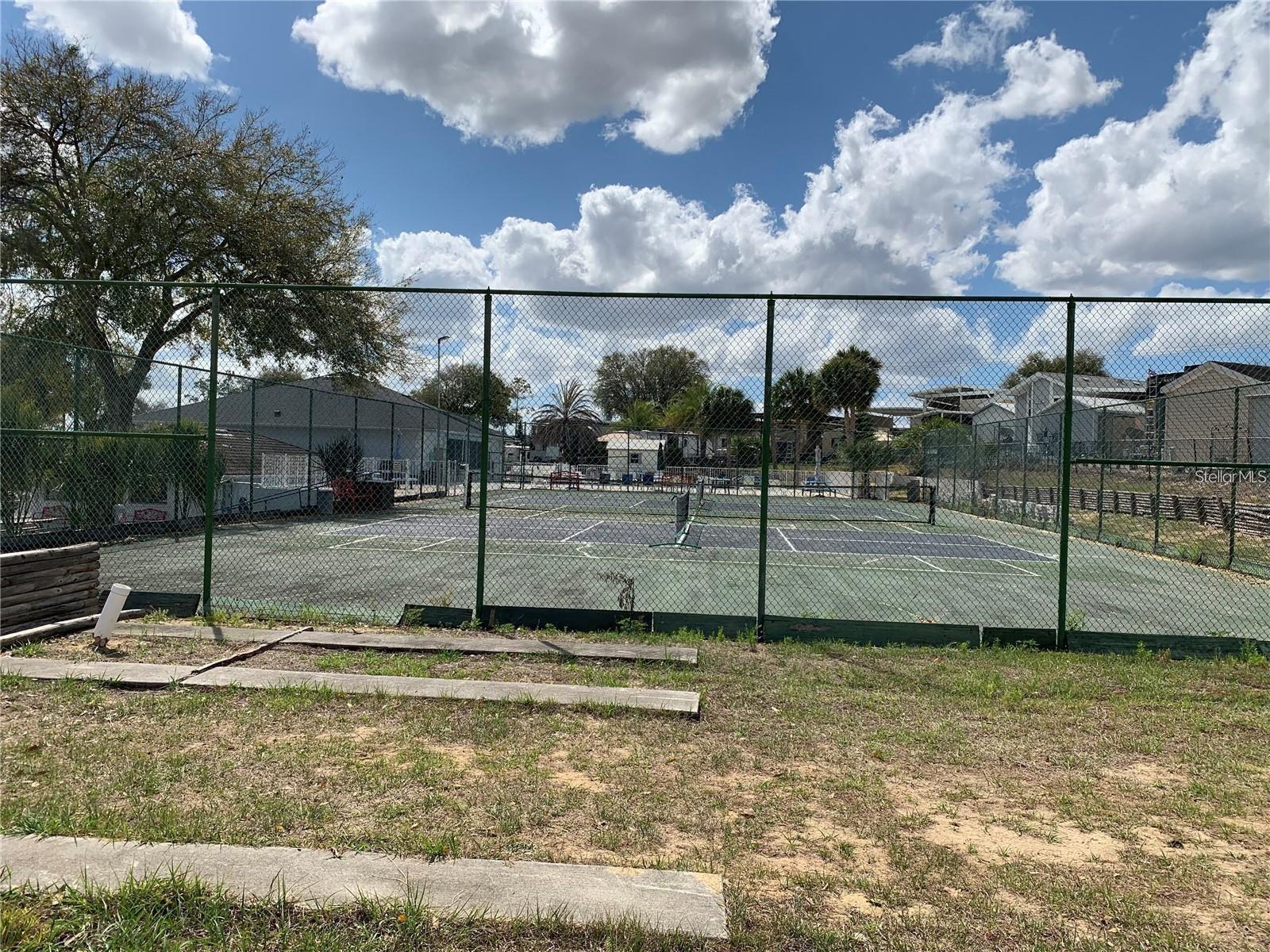 Fenced outdoor tennis court with community buildings and palm trees beyond it.