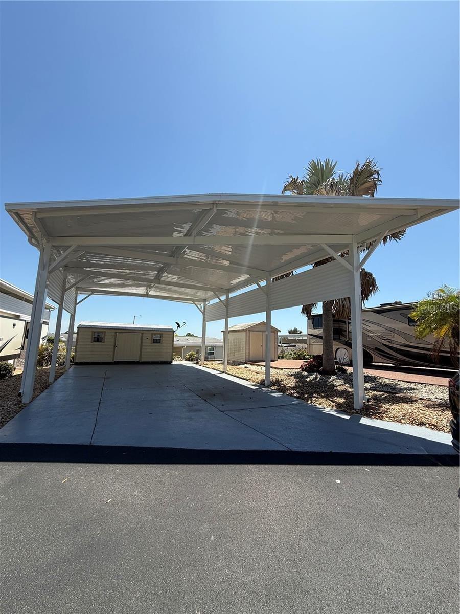 Covered carport over a paved parking area, with a storage shed and palm tree nearby.