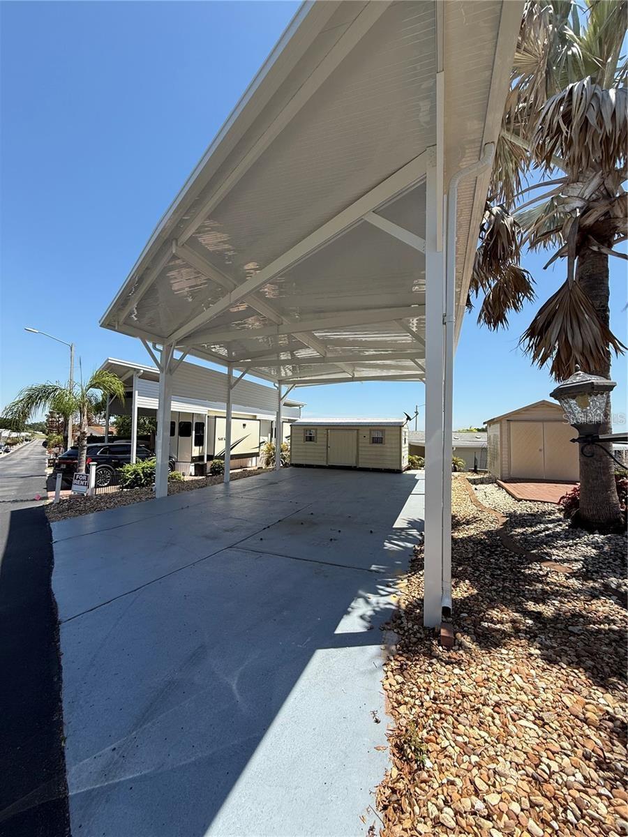Covered carport with a wide concrete parking pad beside manufactured homes and a palm tree.