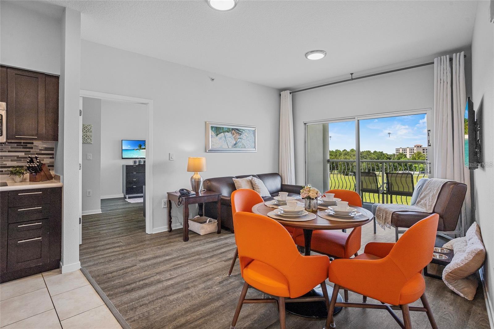 Open living and dining area with orange chairs and sliding doors to a balcony overlooking trees and lawn.