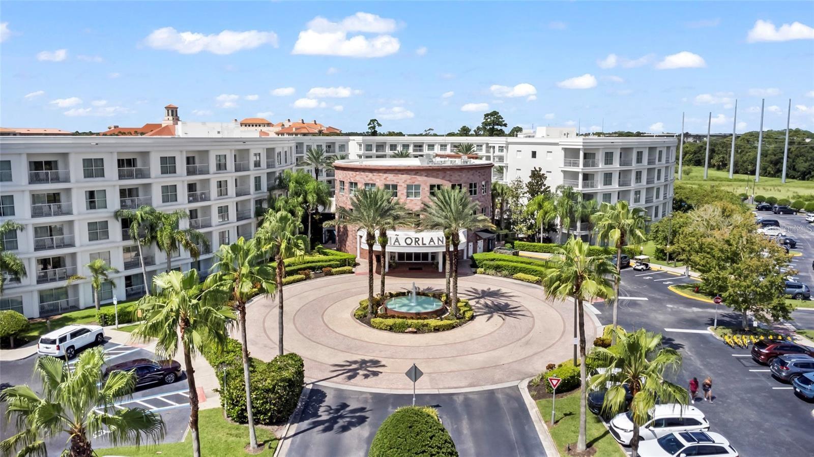 Aerial view of a condominium complex with a circular fountain driveway and palm-lined entrance.