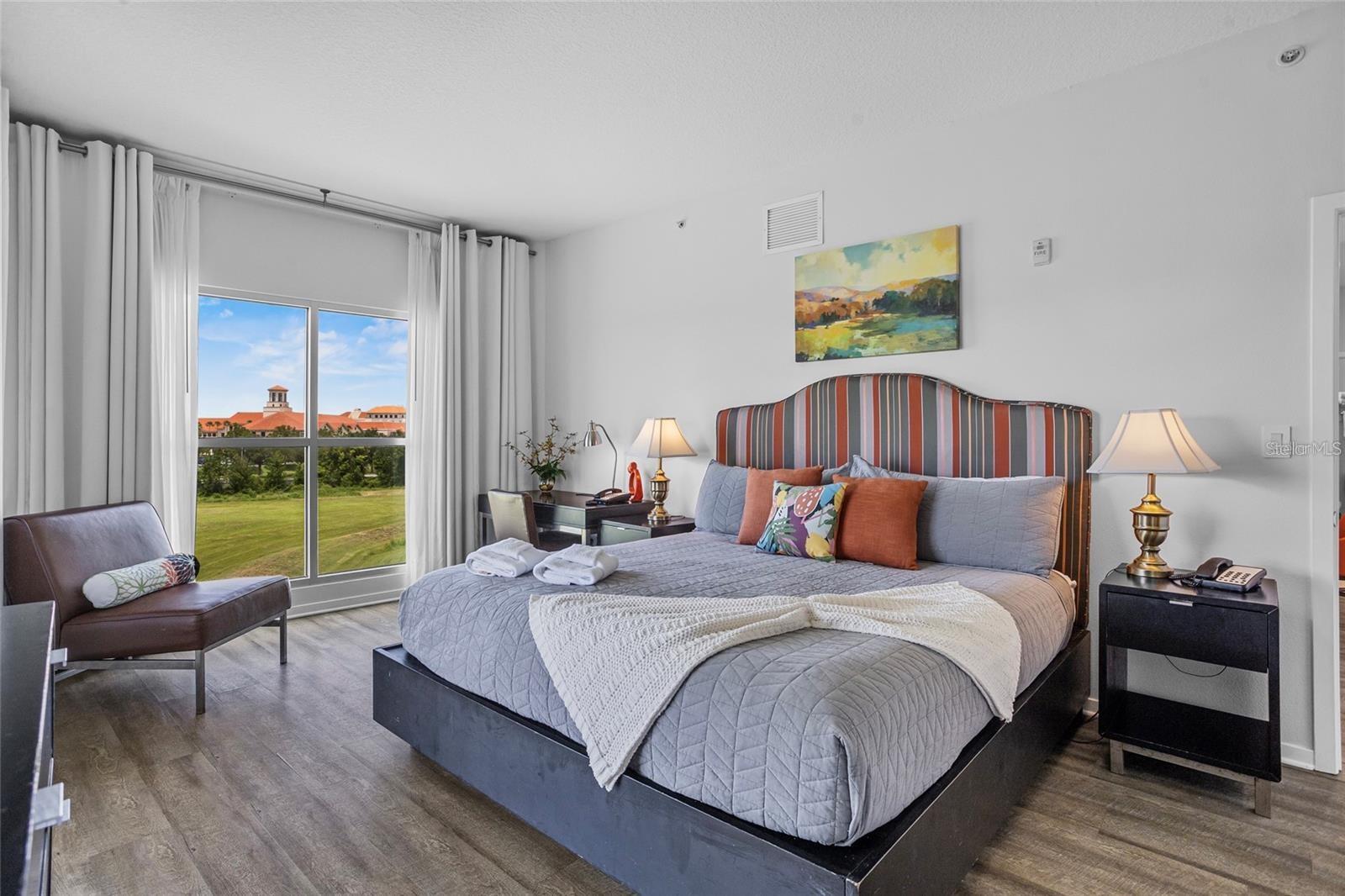 Furnished bedroom with a large gray bed and a window overlooking green grounds and red-roofed buildings.