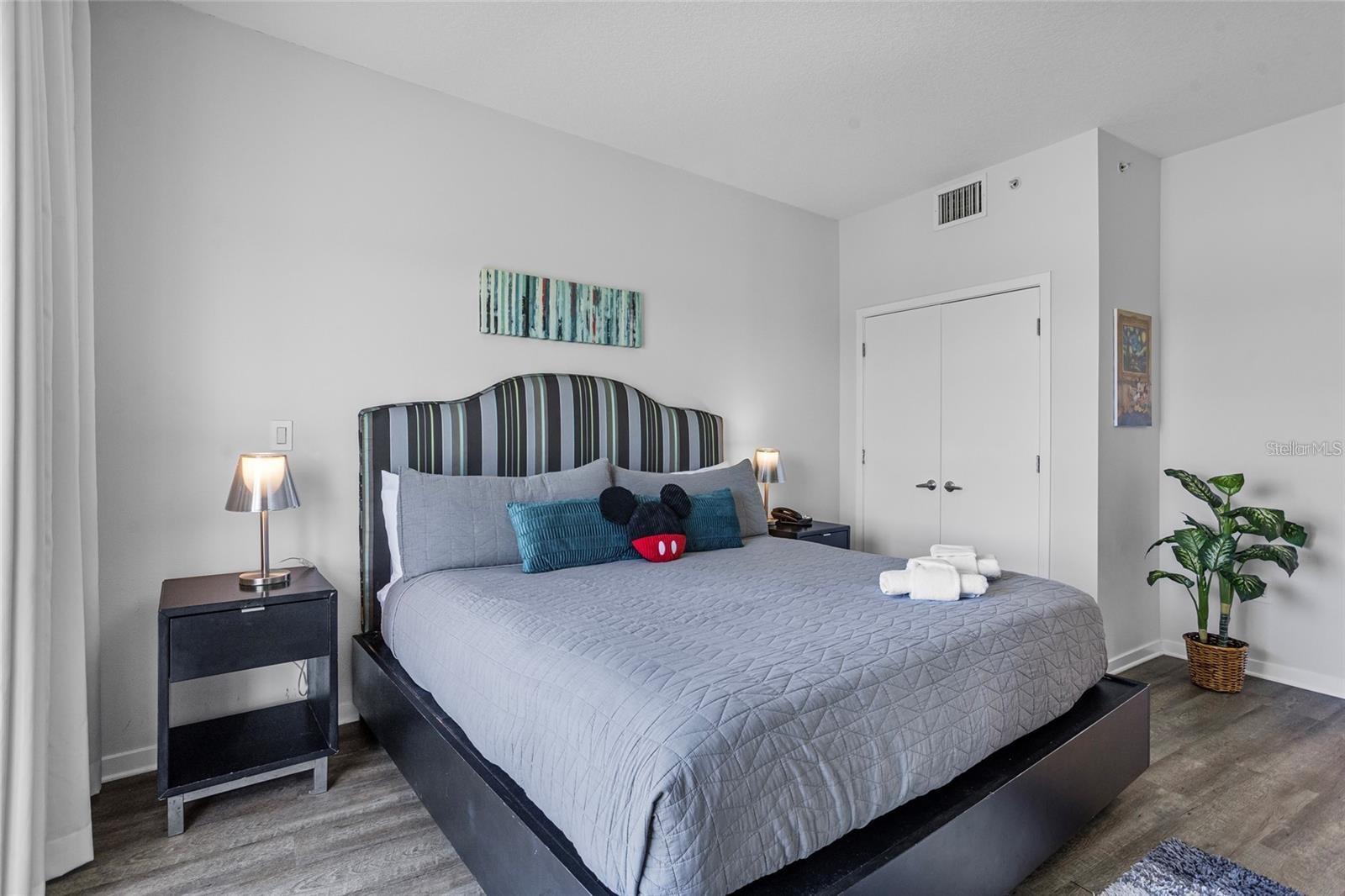 Bedroom with a large gray bed, striped headboard, bedside lamps, and a potted plant.