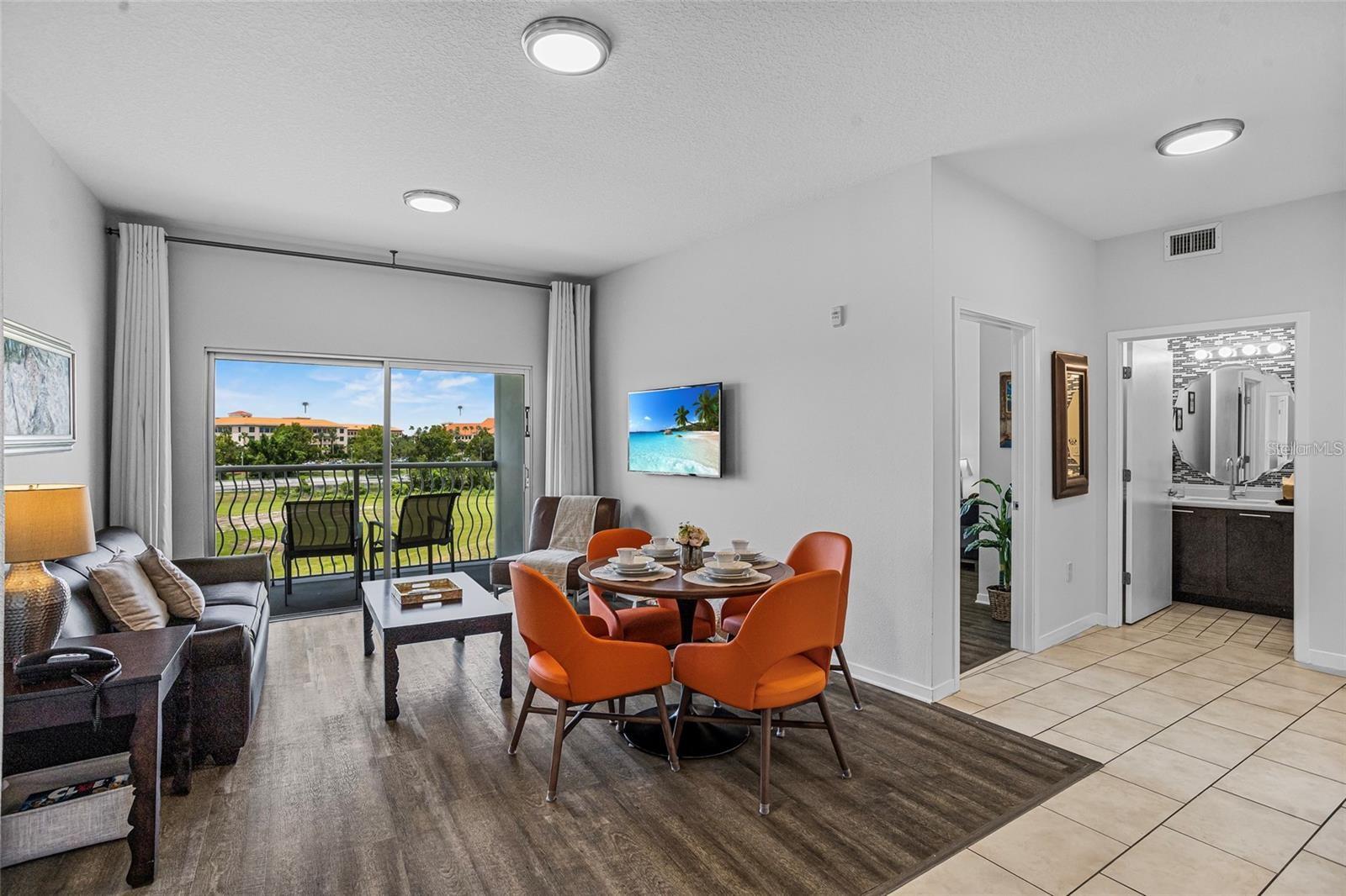 Living and dining area with orange chairs and sliding doors to a balcony overlooking landscaped grounds.