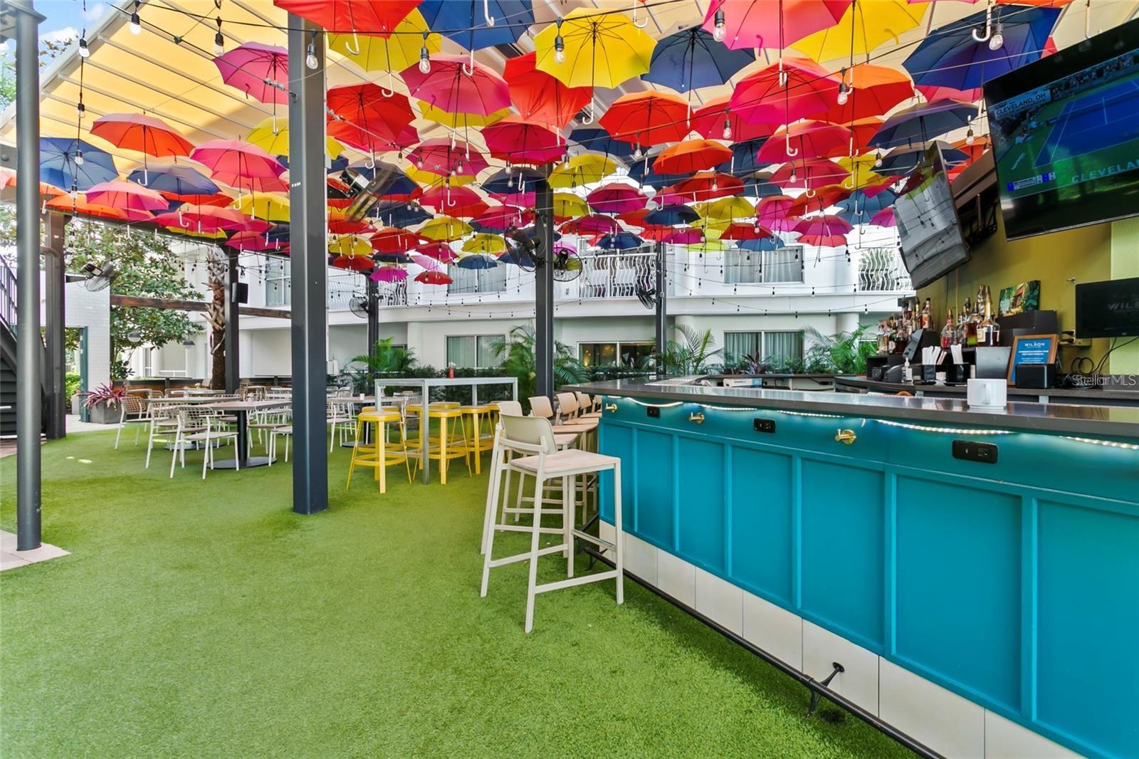 Covered courtyard bar with a turquoise counter and colorful umbrellas suspended overhead.