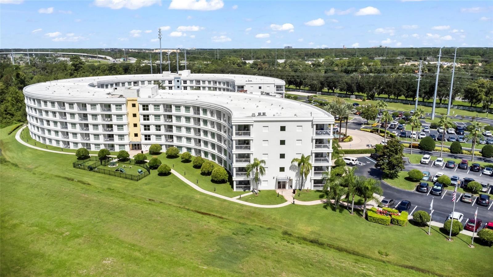 Curved white residential building with landscaped grounds and parking lots, viewed from above.