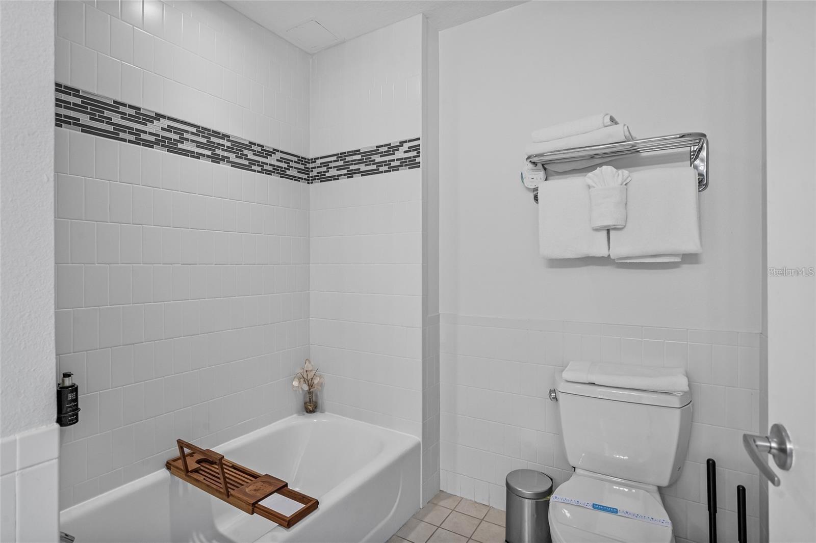 Bathroom with a white bathtub, wooden tray, tiled accent stripe, and toilet.