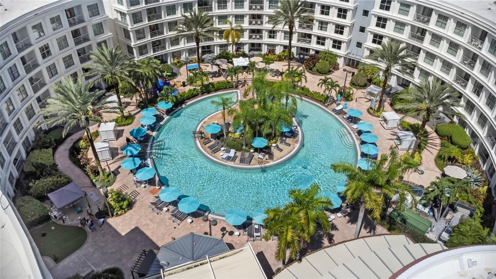 Aerial view of a courtyard pool with a palm-lined island and turquoise umbrellas.