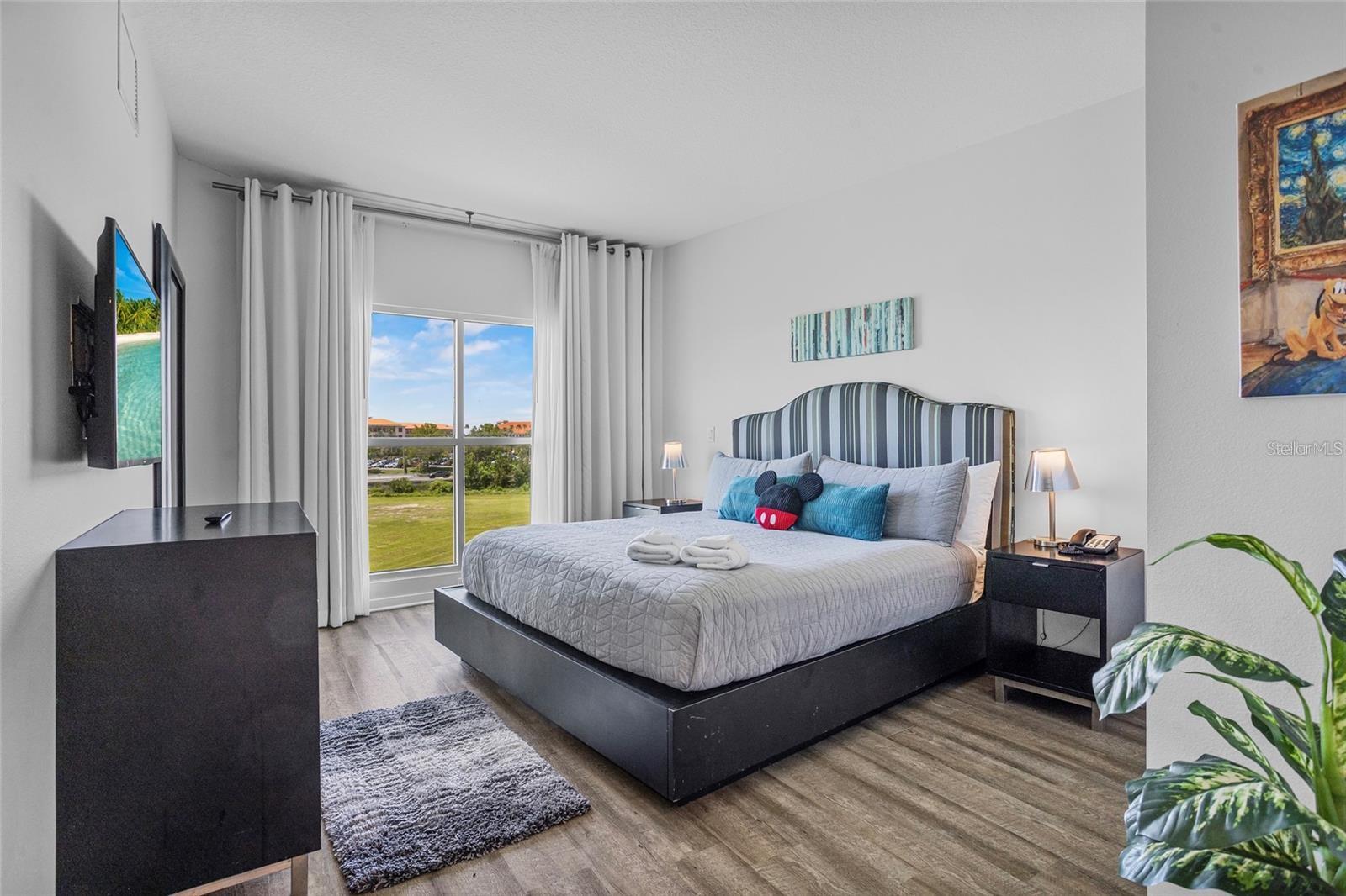 Furnished bedroom with a king bed, striped headboard, and large window overlooking green grounds.