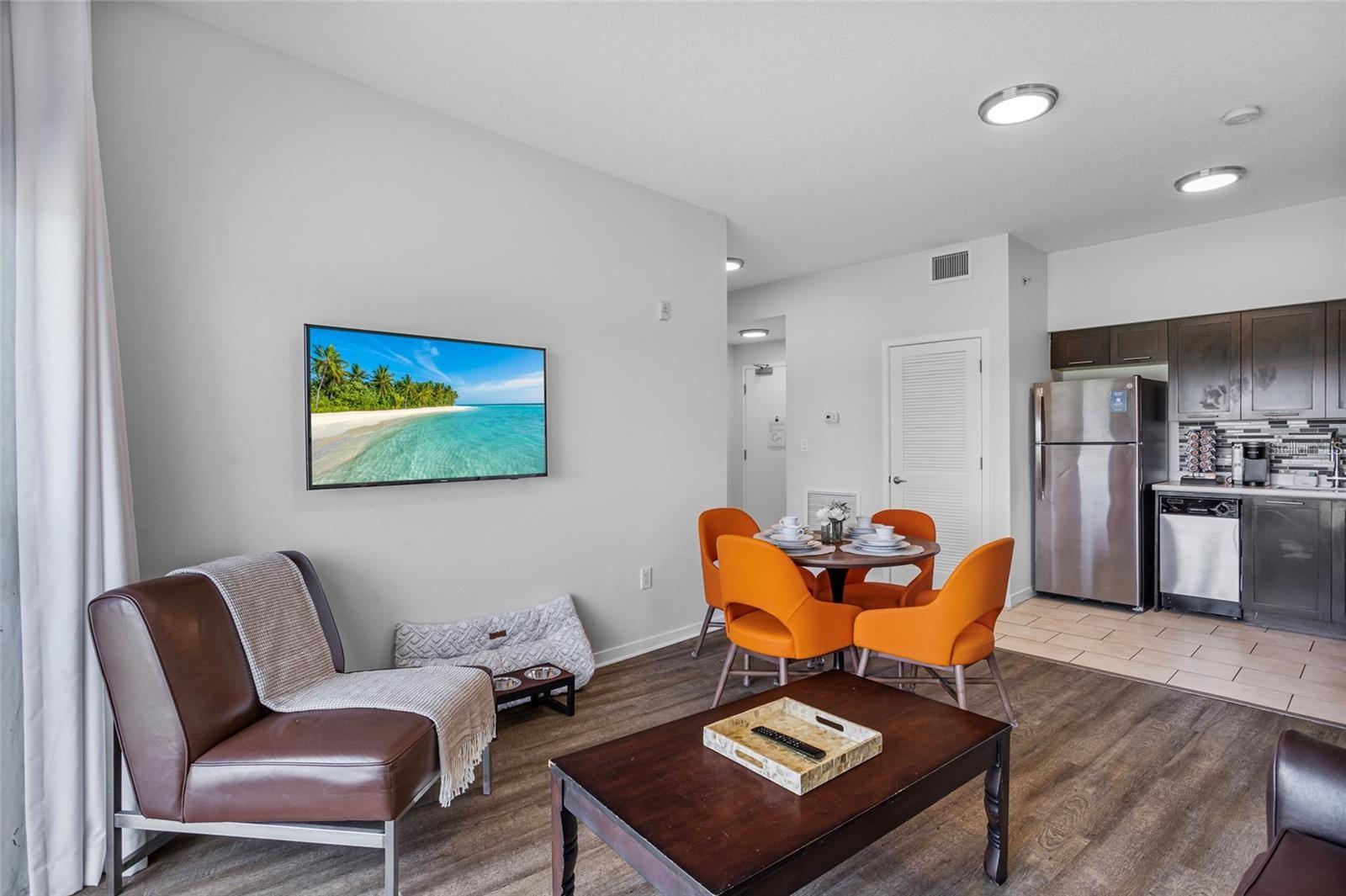 Open-plan condo living and dining area with orange chairs, brown leather seating, and a dark-cabinet kitchen.