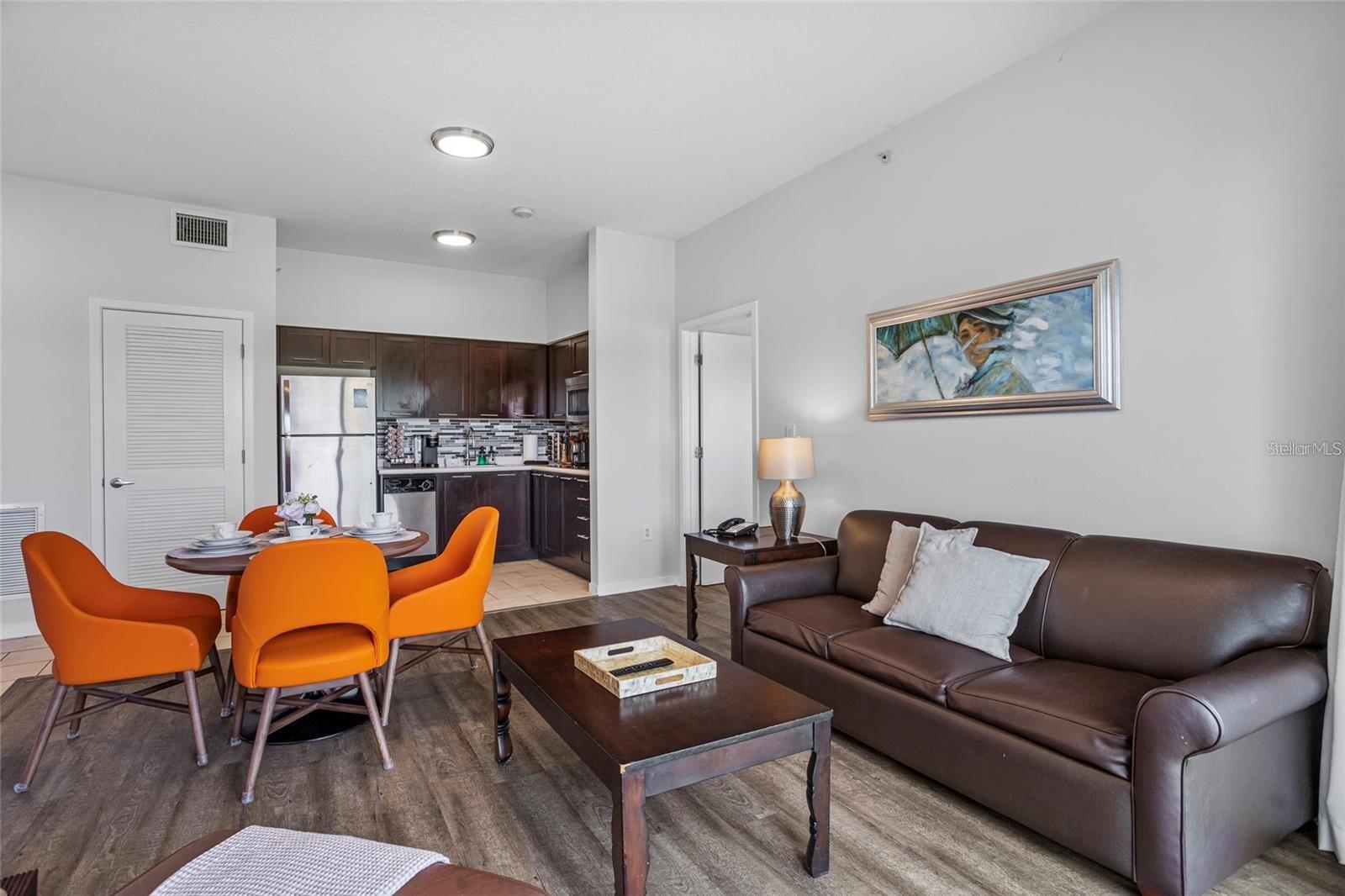 Furnished living room with brown leather sofa, orange dining chairs, and an open kitchen.